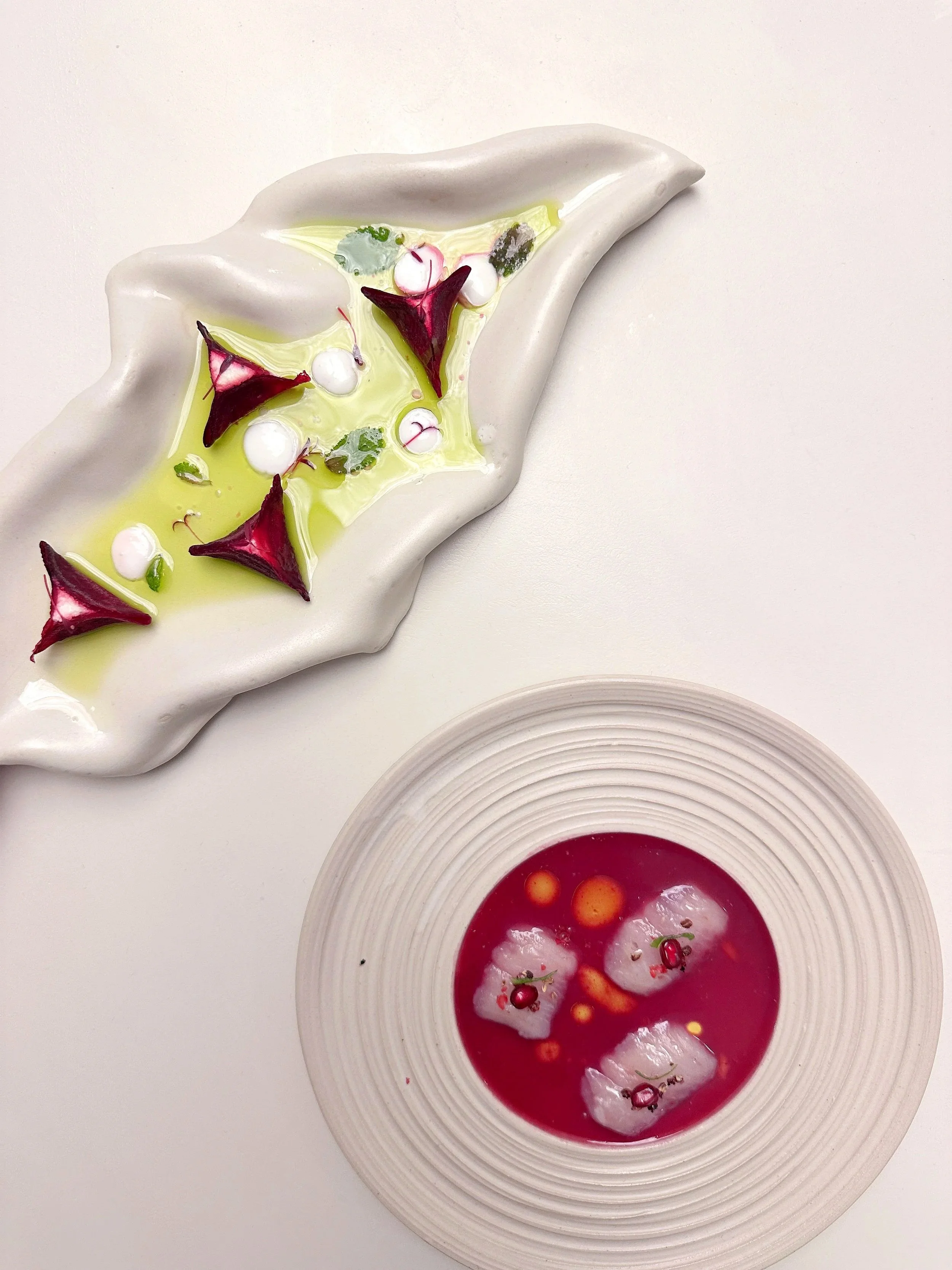 Une assiette blanche avec un plat décoratif comprenant des éléments verts et violets, et une coupe blanche contenant une soupe rouge avec des fruits ou légumes flottants et des garnitures.