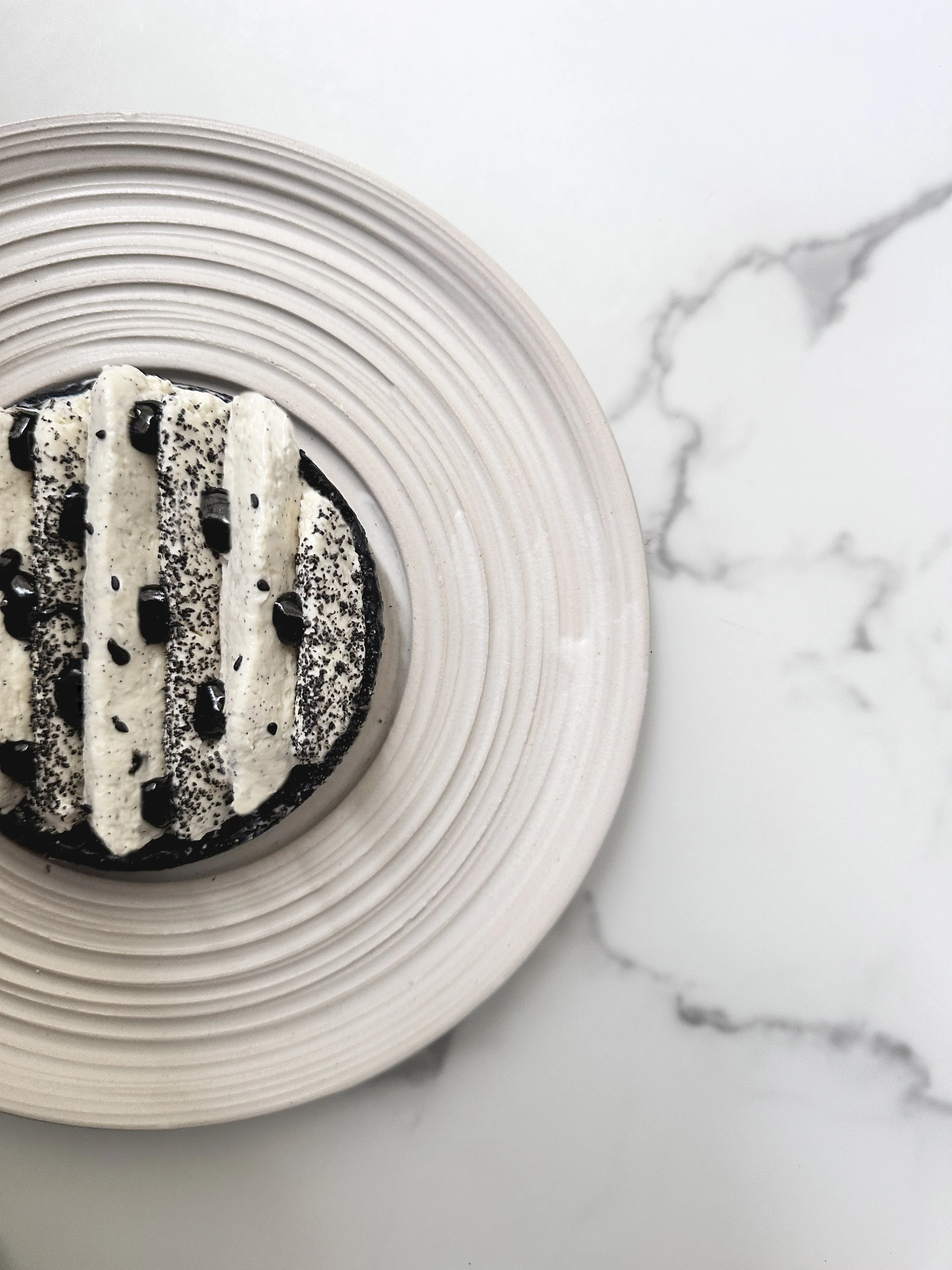 Tartelette vanille sésame noir présentée sur une assiette blanche à cercles concentriques, sur une surface en marbre blanc avec veines grises.