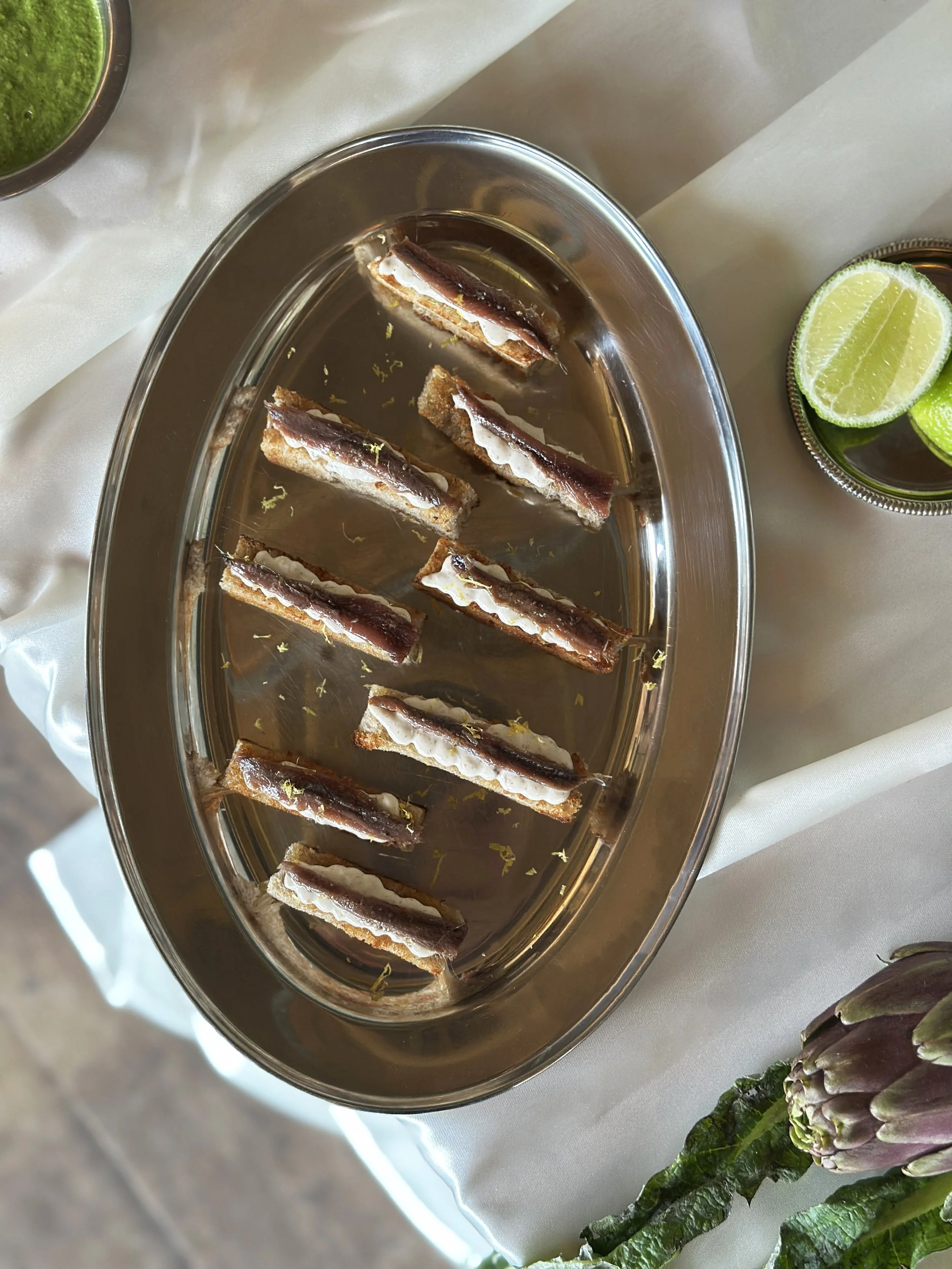 Toasts de ricotta anchois et zeste de citron, servis sur un plat en acier inoxydable, accompagnés de quartiers de citron vert, posés sur une nappe blanche.