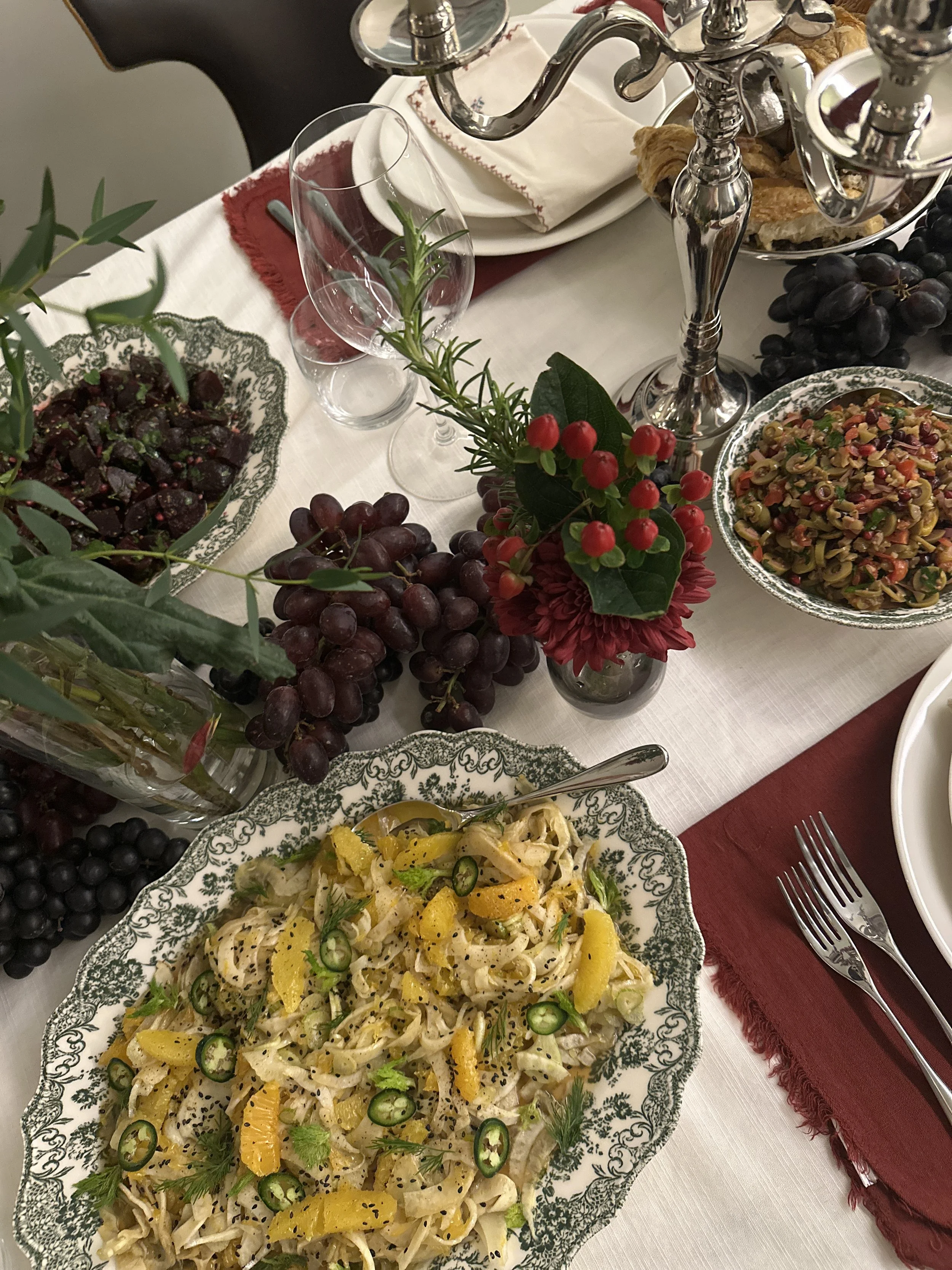 Une table de fête avec un plat de pâtes, un bol de salade de fenouil, un bol de salade d'olives, une caraffe de vin, une jolie centerpiece et des couverts. La table est décorée pour un repas élégant.