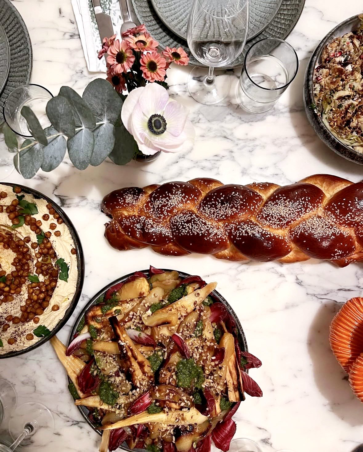 Repas de fête avec un pain brioché glacé, un plat de houmous, une salade de légumes, des plats de fête et des verres à vin sur une table en marbre.