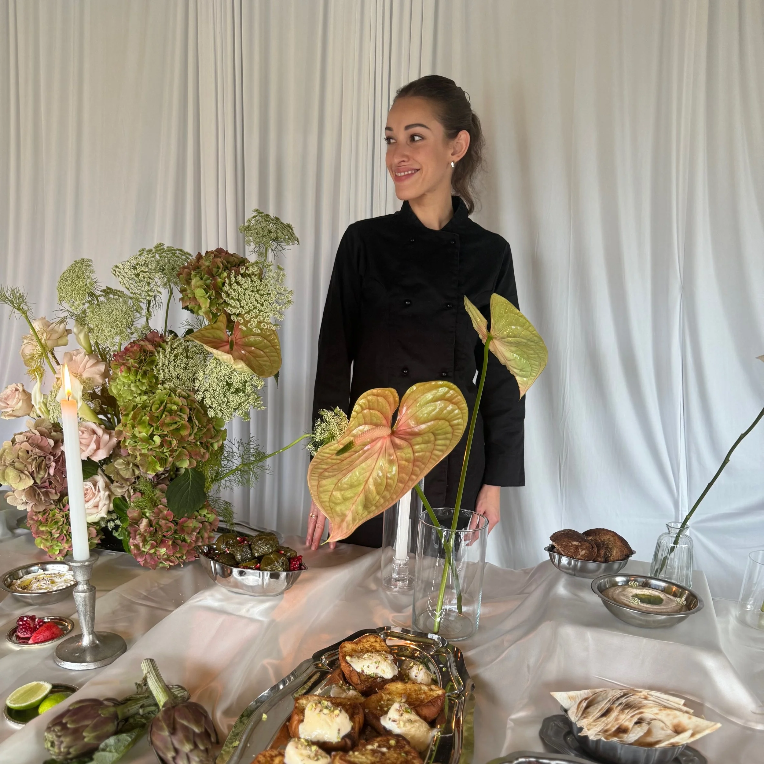 Une femme souriante portant une veste noire, se tient devant une table de pâtisseries et de fleurs, avec un fond de rideaux blancs.