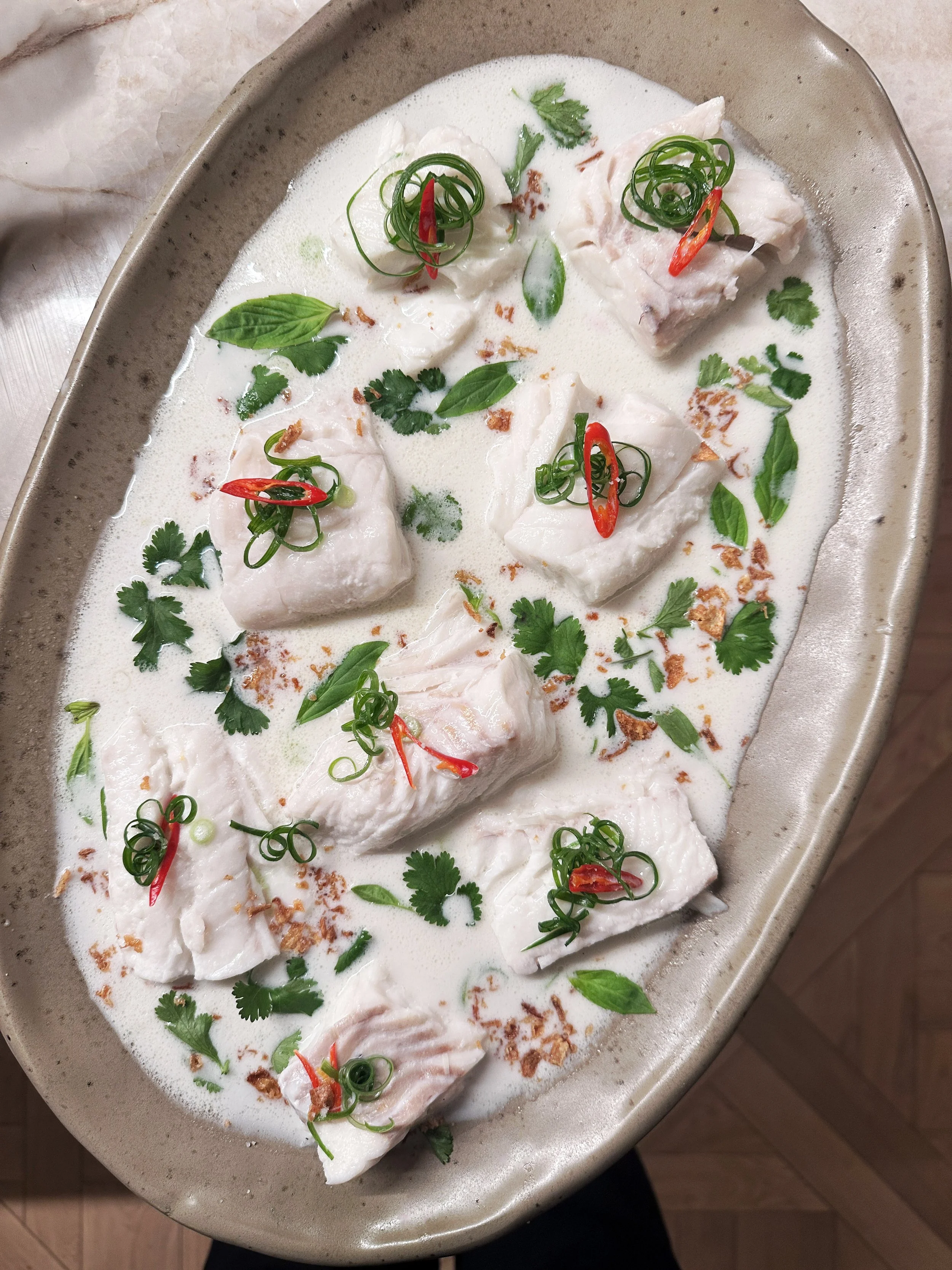 Poisson blanc en sauce au lait de coco, garni de coriandre, basilic, piment rouge et oignons verts, présenté dans un plat en céramique