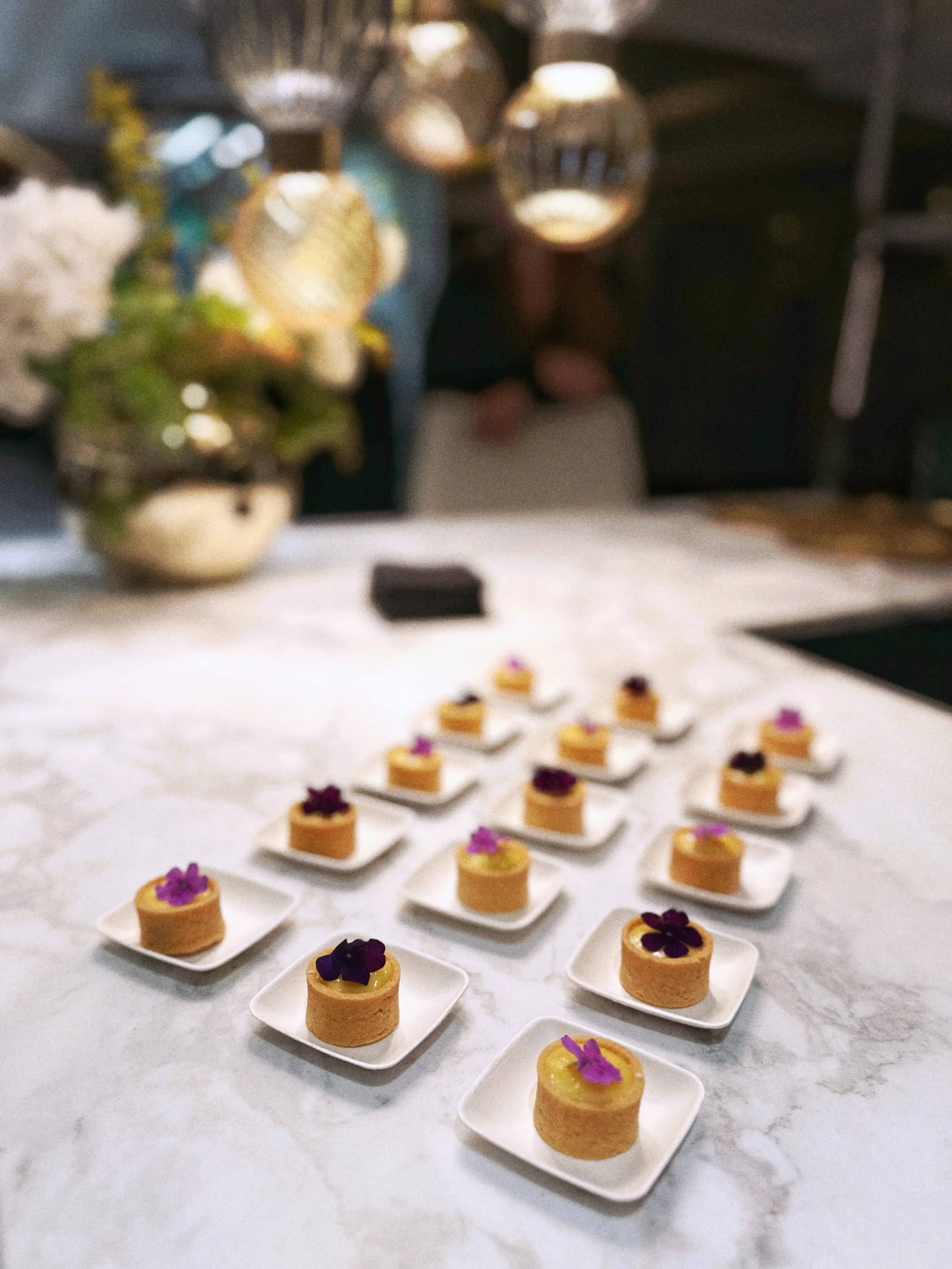 Une table avec plusieurs petites desserts présentés sur des assiettes blanches, décorés de fleurs violettes, et en arrière-plan, un vase avec des fleurs blanches et un décor lumineux. La photo est floue.