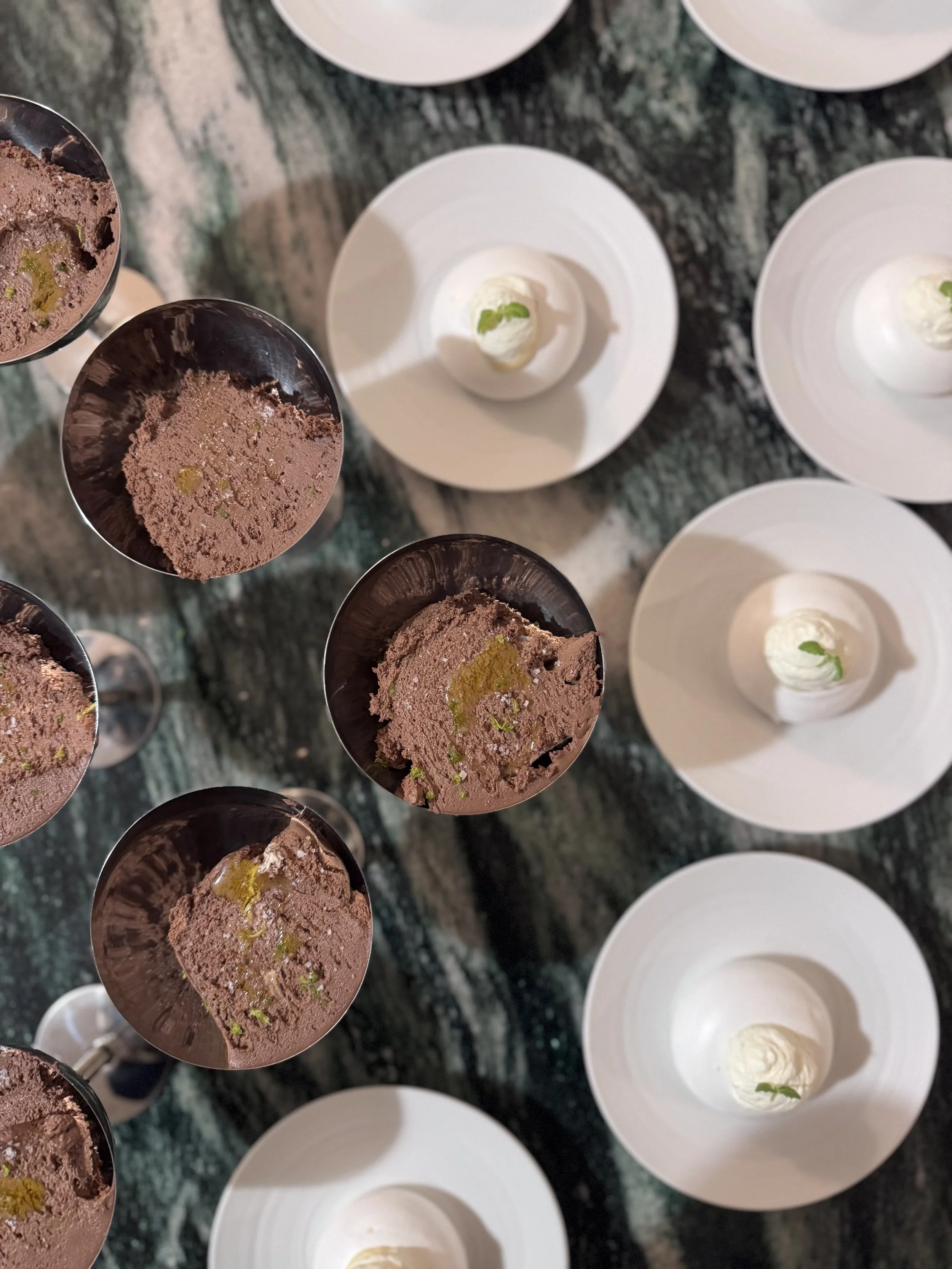 Gobelets de mousse au chocolat avec des zestes de citron vert, entourés d'assiettes blanches avec une portion de crème ou de sorbet à la menthe, sur une table en marbre.