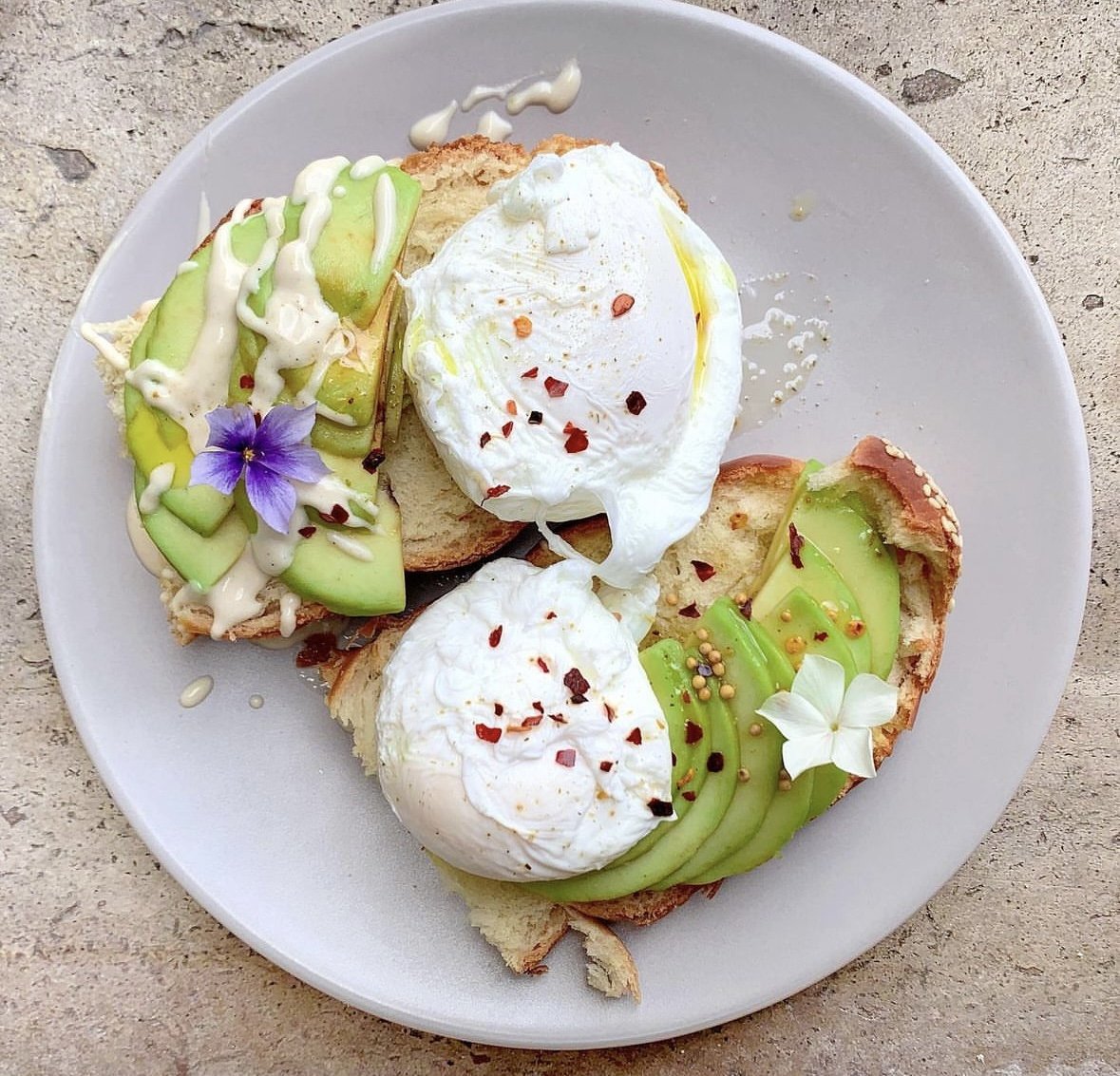 Tartines de pain avec avocat et oeufs pochés, décorées de fleurs comestibles et de piment rouge, servies sur une assiette blanche.