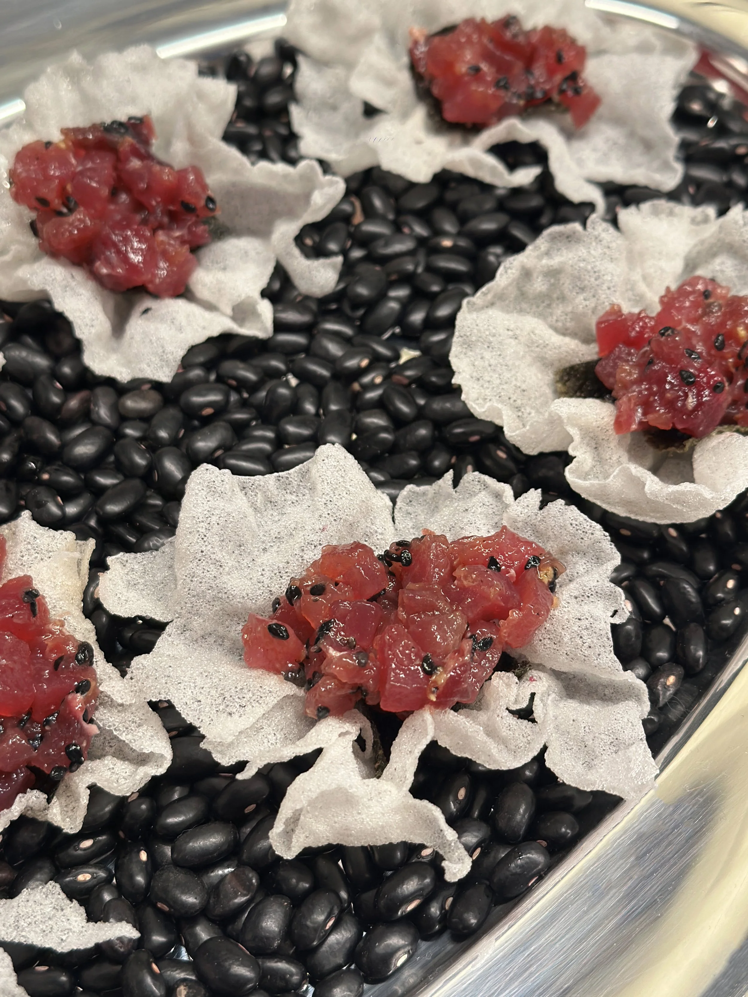 Image montrant des flocons de papier de riz garnis de morceaux de tartare de thon avec des graines de sésame noires, sur un fond de graines de sésame noires