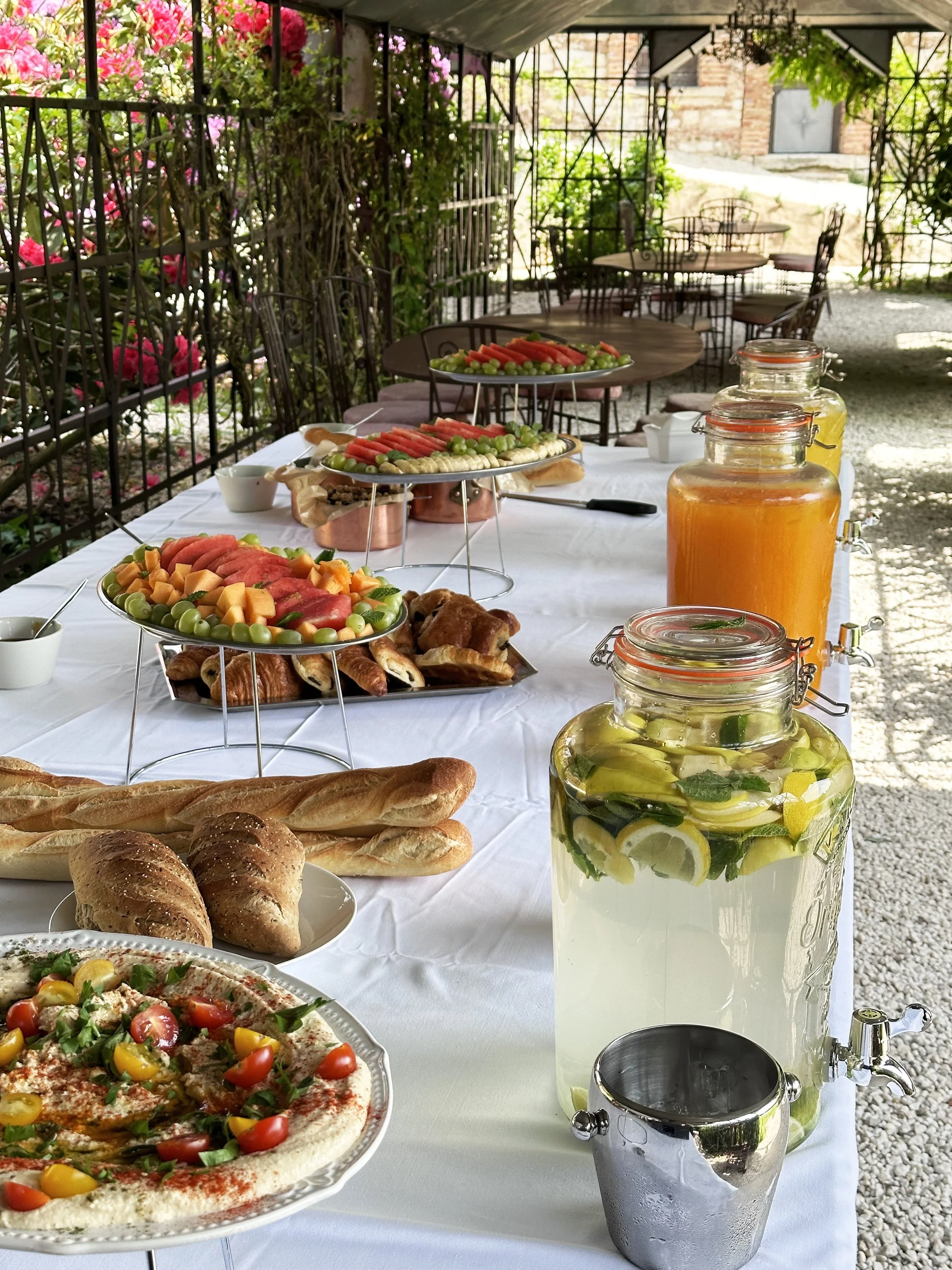 Buffet avec des fruits, boissons, pain, houmous, fruits et autres mets, en plein air sous un abri avec des chaises et une végétation fleurie en arrière-plan.
