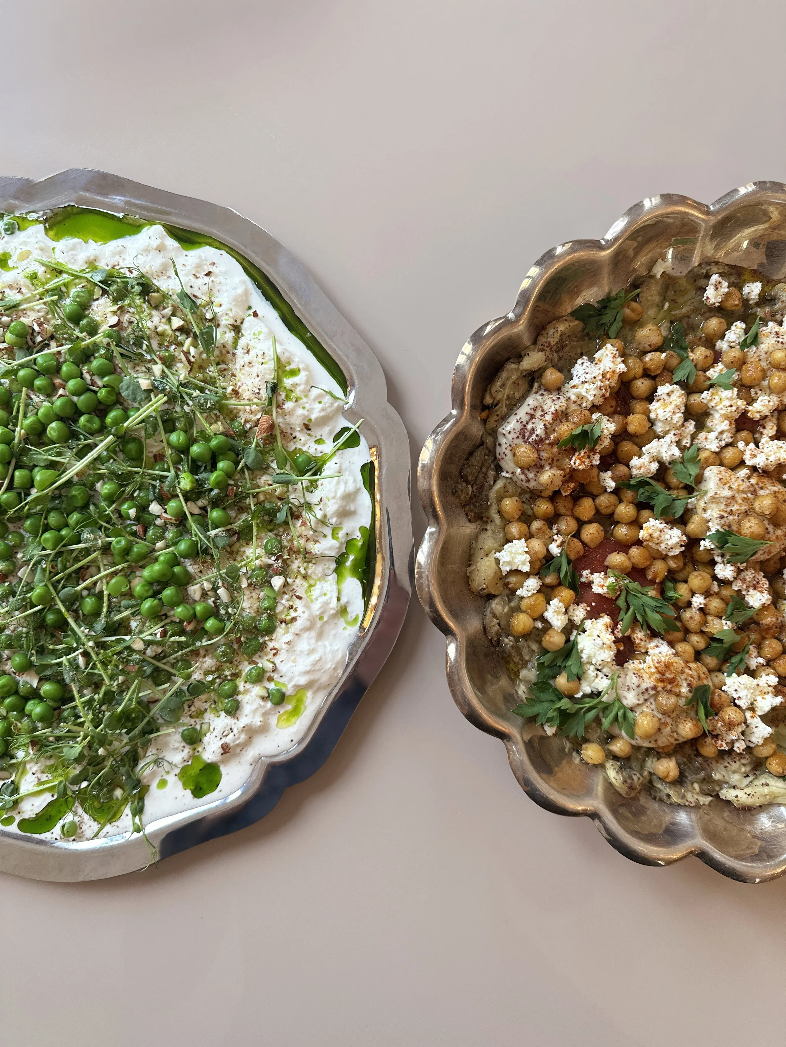 Deux plats de cuisine méditerranéenne, l'un avec de la burrata, des petit pois frais, amande grillée, huile verte et des pousses de pois, l'autre avec des pois chiches, aubergine, tomates, feta, des herbes et du paprika dans un plat en argent.