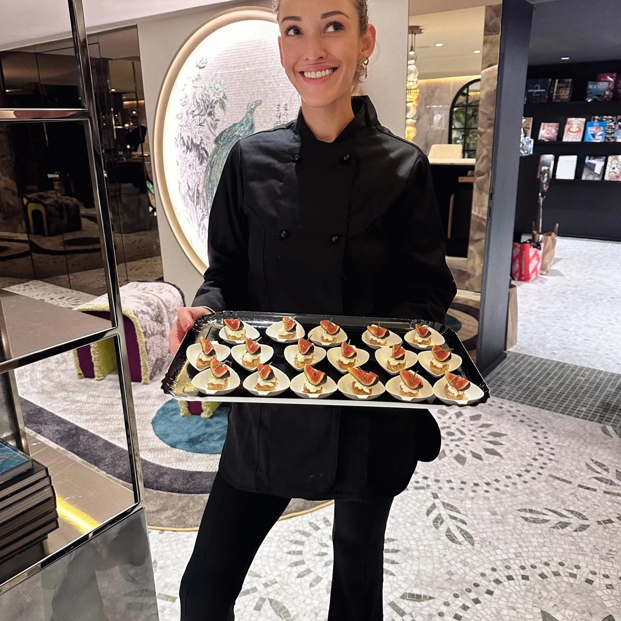 Une femme en uniforme noir sourit en tenant un plateau de amuse-bouches dans un décor intérieur moderne et élégant.