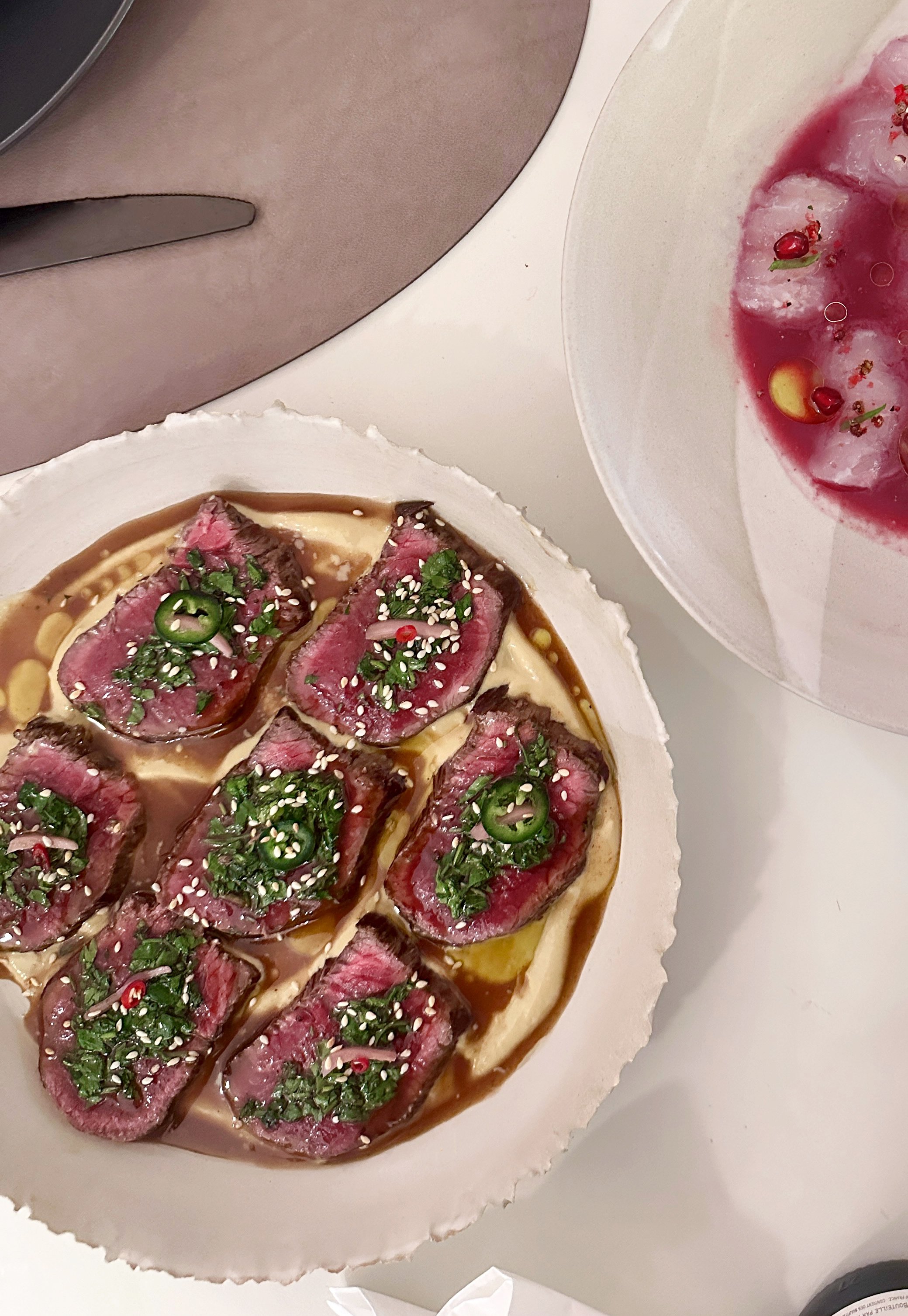 Tataki de bœuf garni de cébettes, graines de sésame, piment jalapeño, et sauce tahini, présentée sur une assiette en céramique, avec un plat de salade contenant des sashimi de daurade et une sauce à la grande