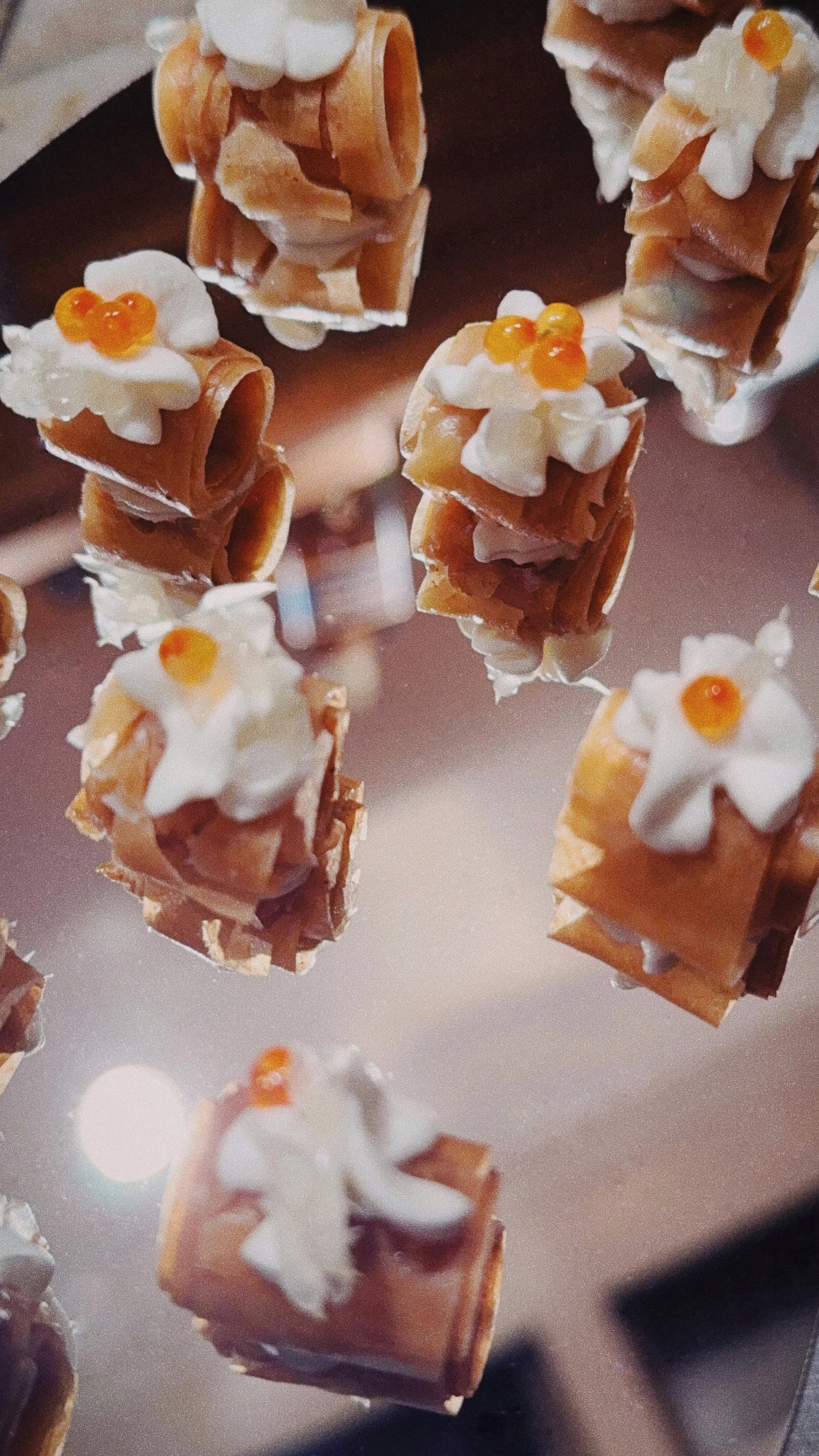 Petites bouchées avec des rouleaux de caramel, garnies de crème et de petites billes orange, présentées sur une surface en miroir.