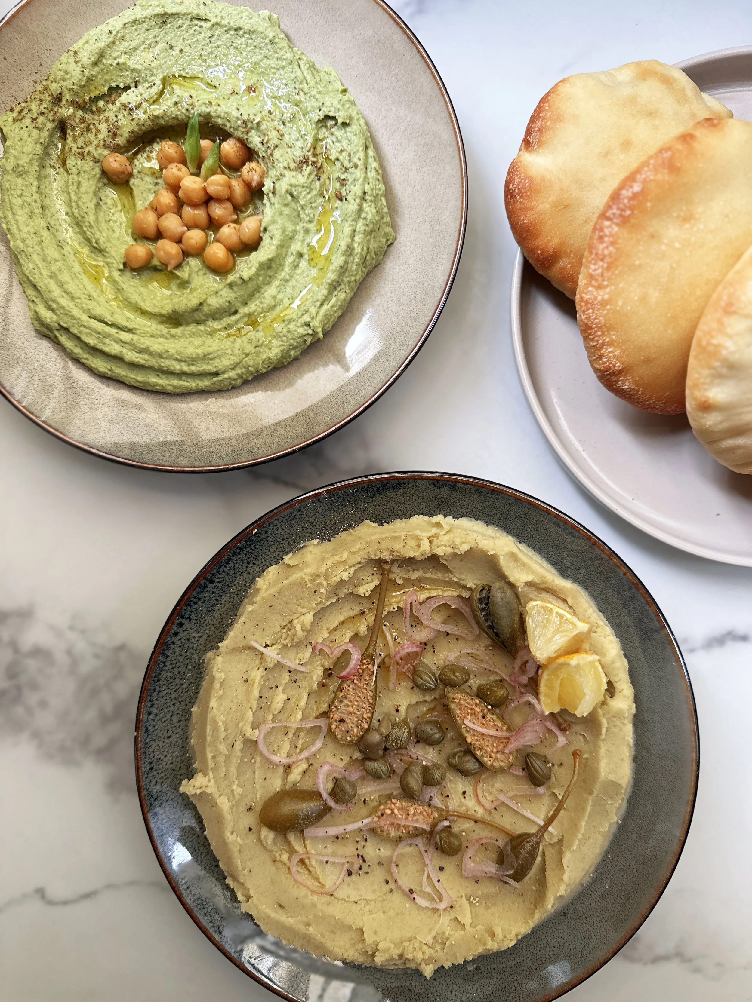 Hummus à l'ail des ours et fava garnie de câpres, oignon, citron, et graines, présenté dans un bol, avec du pain pita à côté.