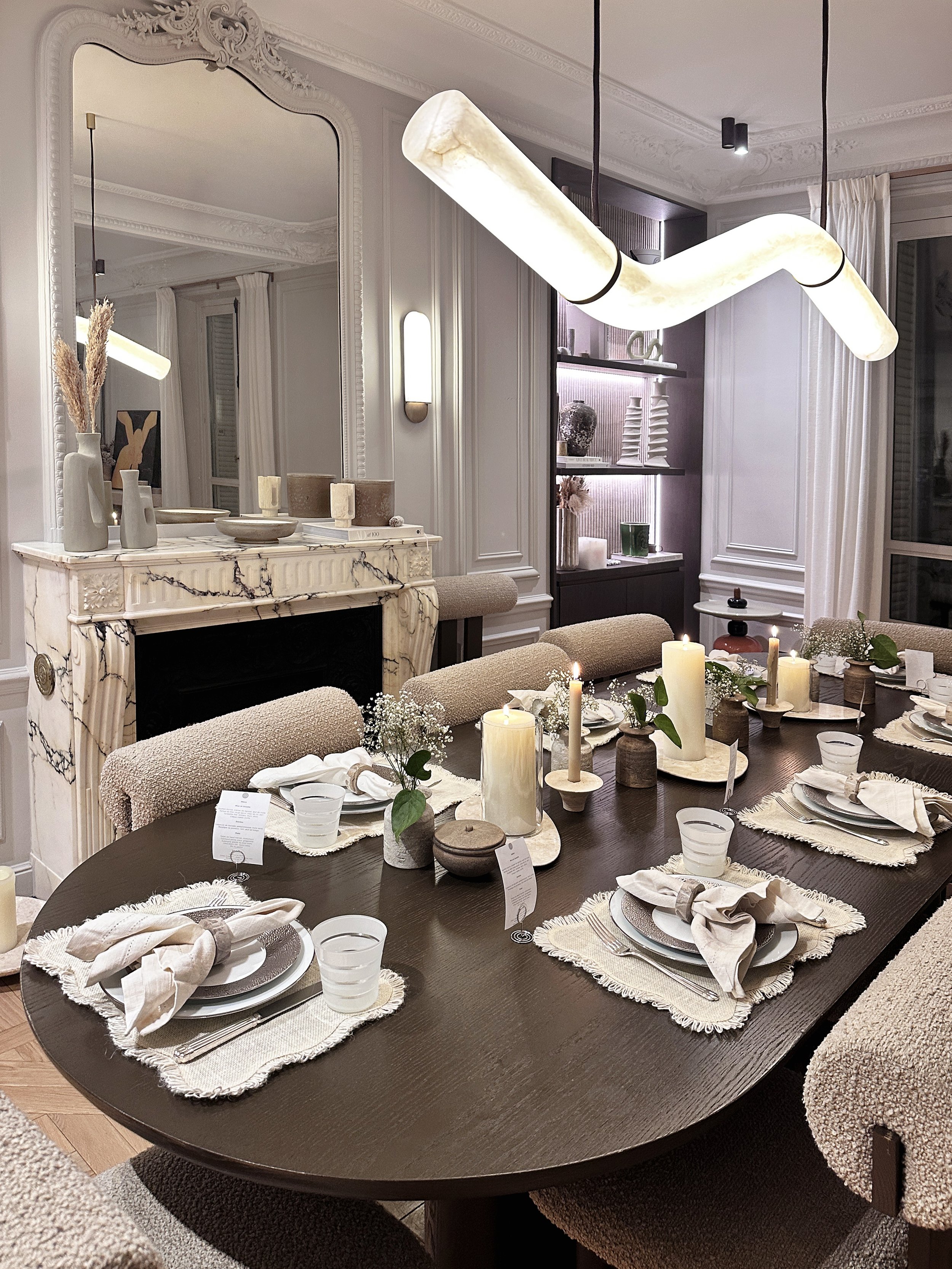 Une salle à manger élégante avec une grande table ronde en bois sombre, des assiettes blanches, des verres d'eau, des serviettes beige pliées et des bougies. En arrière-plan, un manteau en marbre blanc avec des vases et objets décoratifs, un grand mi