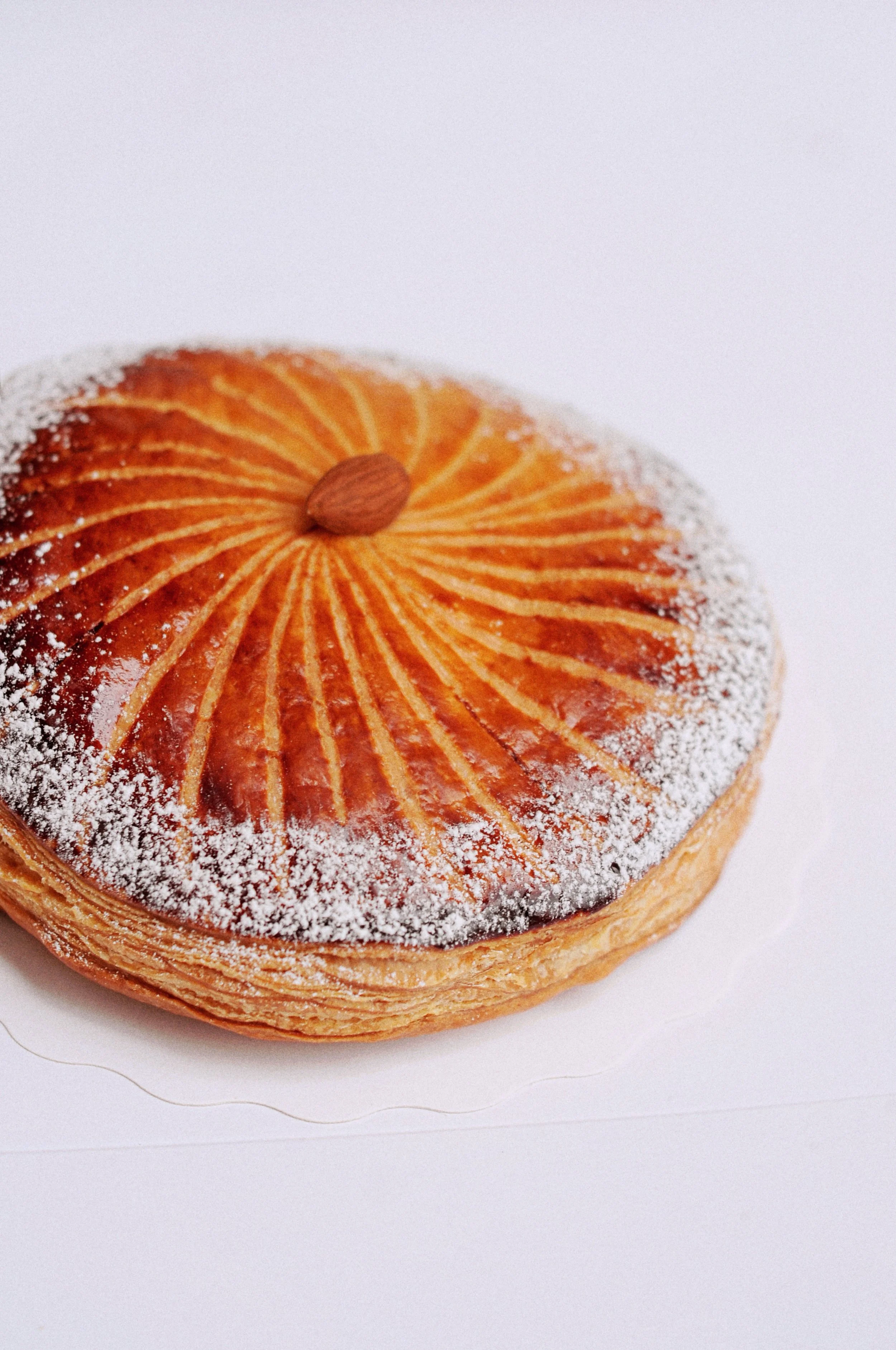 Une galette des rois avec un motif rayonnant sur la surface, saupoudrée de sucre glace, sur un fond blanc.