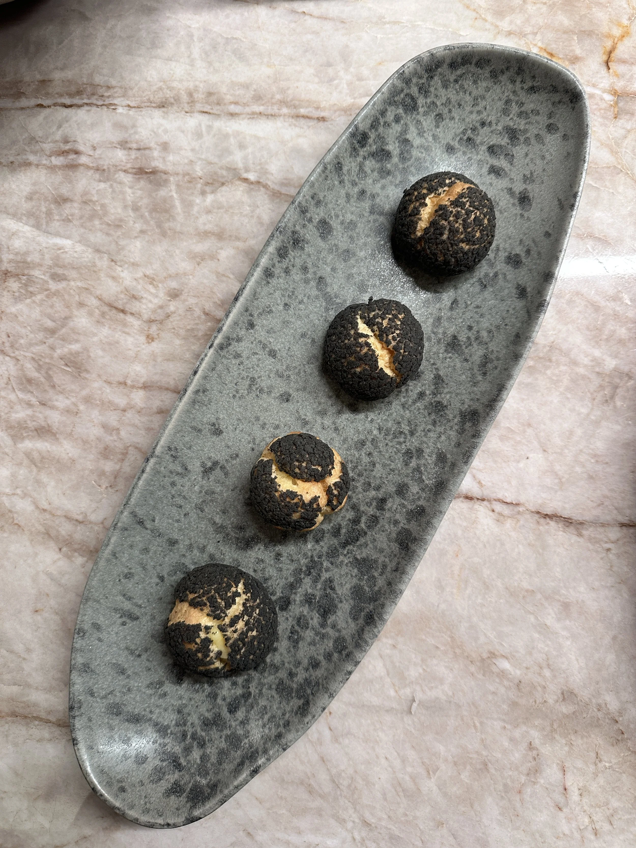 Choux craquelin vanille et sésame noir sur une assiette rectangulaire grise, sur une surface de marbre clair.