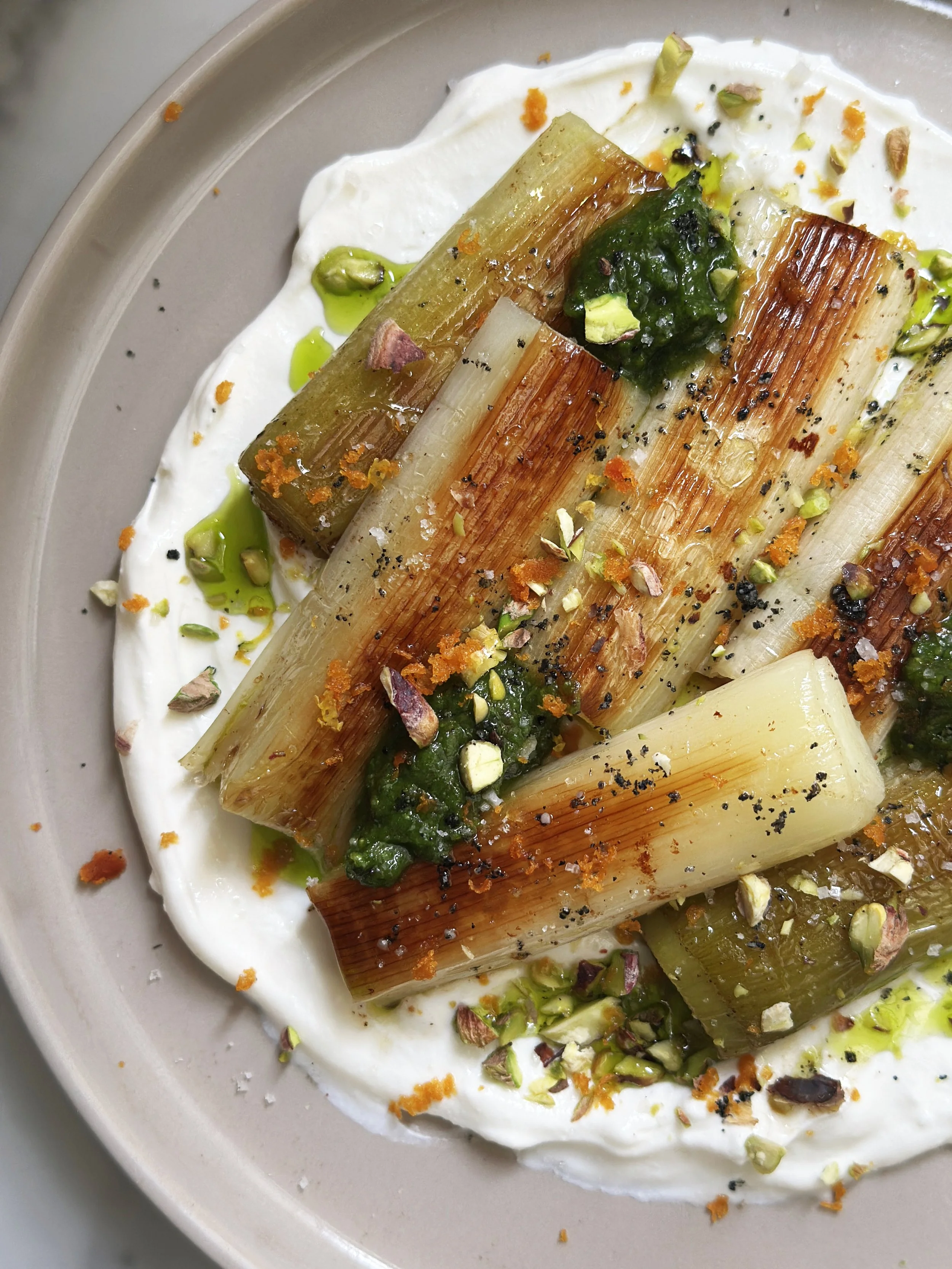 Poireaux grillés avec pistaches, labneh fumé, huile verte de poireau, pesto de pistache, boutargue, épices et zestes d'orange, présentés dans une assiette.
