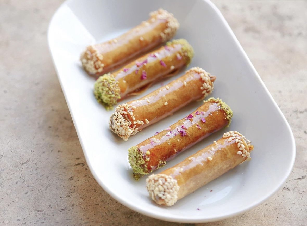 Six cigares aux amandes et pistaches décorés avec des graines de sésame, de pistache et des fleurs. Présentés dans un plat blanc.
