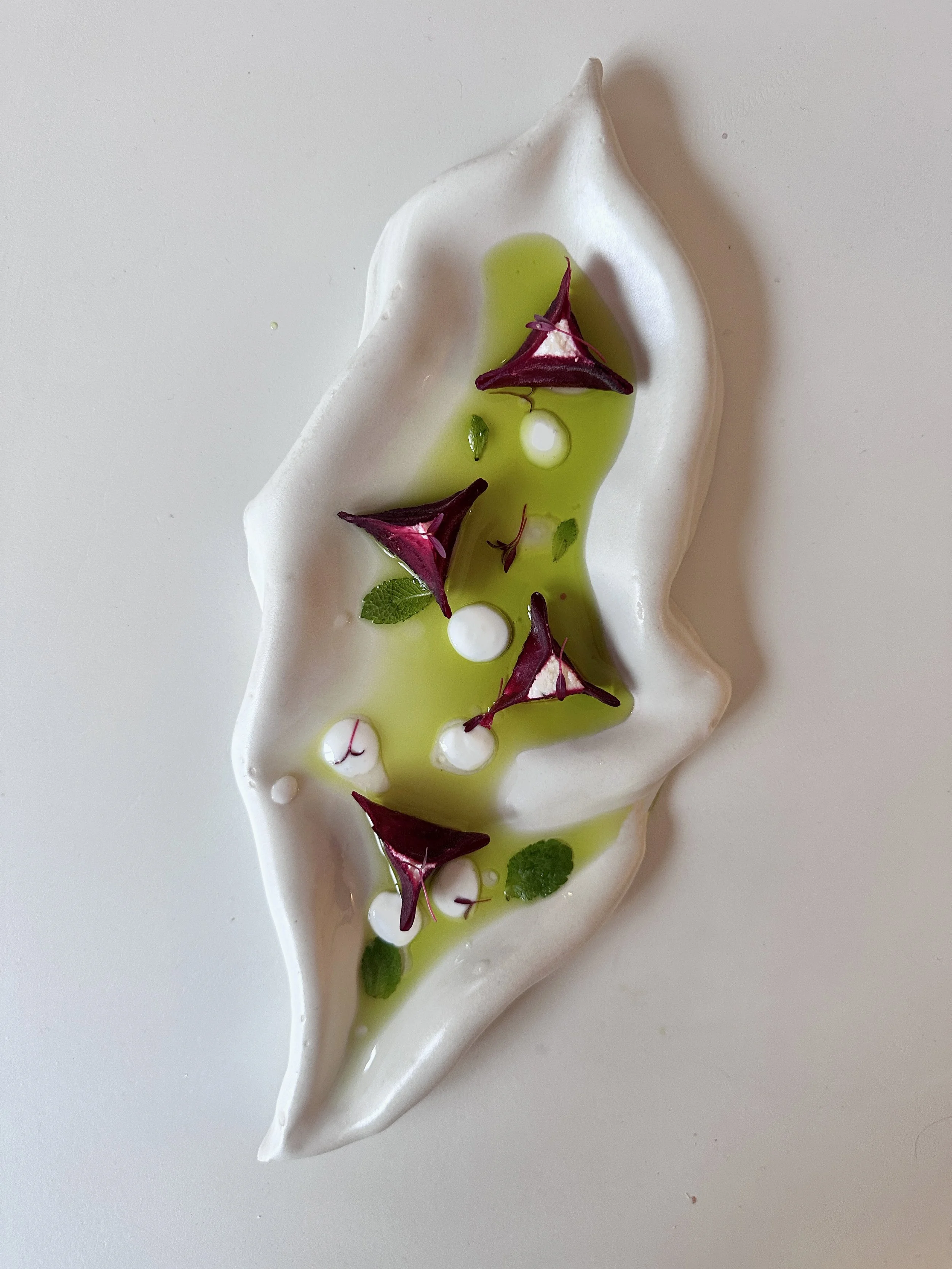 Une présentation culinaire artistique sur une assiette blanche avec une sauce verte, des ravioles de betterave, feuilles de menthe, des gouttes de sauce blanche, et des fleurs comestibles colorées.
