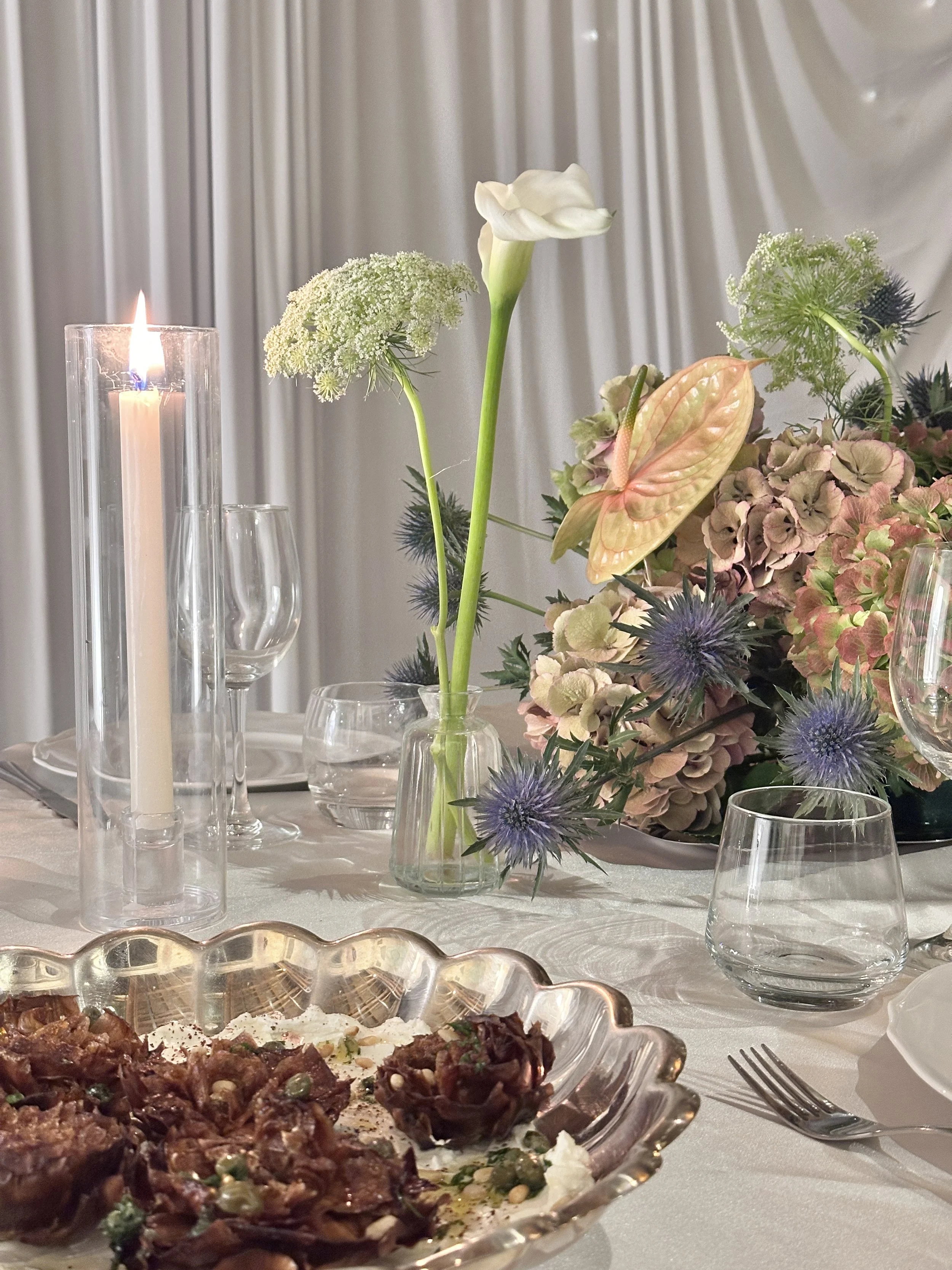 Une table avec un bouquet de fleurs, des bougies, des verres, et un plat de nourriture.