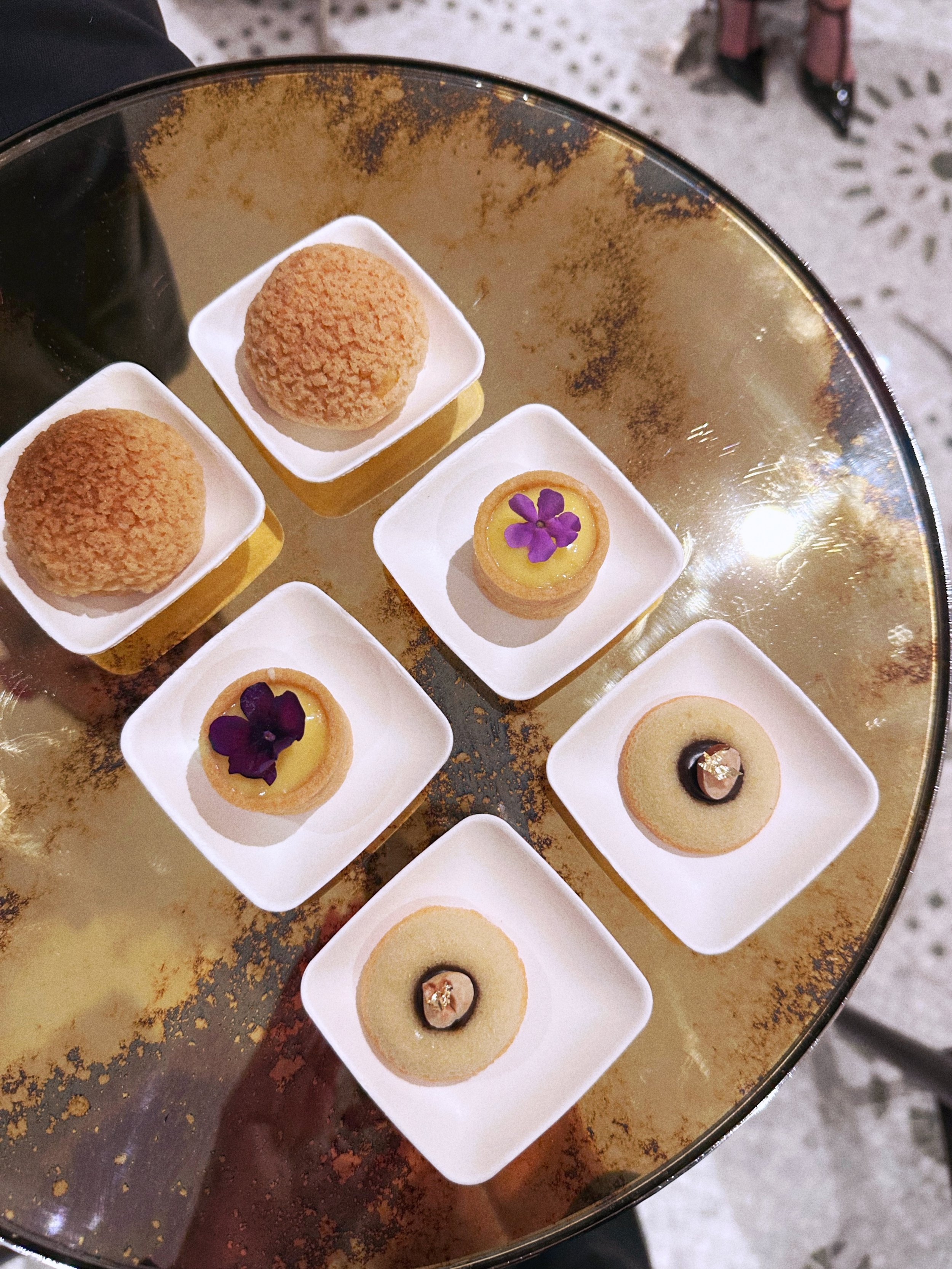 Une assiette en miroir contenant plusieurs petits desserts dans des coupelles blanches. Deux ont un biscuit rond avec une texture croustillante, un autre dessert a une garniture de crème ou de gel avec une petite fleur violette décorative, et d'autre