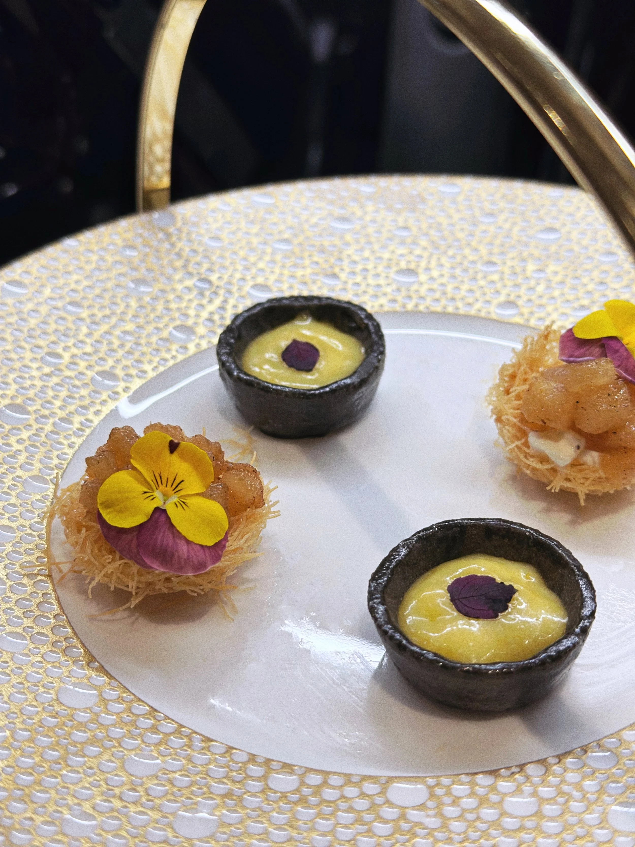 Petites tartelettes en coque au charbon, garnies d'une crème au citron et décorées de pousses de shiso pourpre, et bouchées de kadaïf à la poire rôtie avec pétales de fleurs jaunes, sur une assiette blanche avec un bord en dentelle dorée.