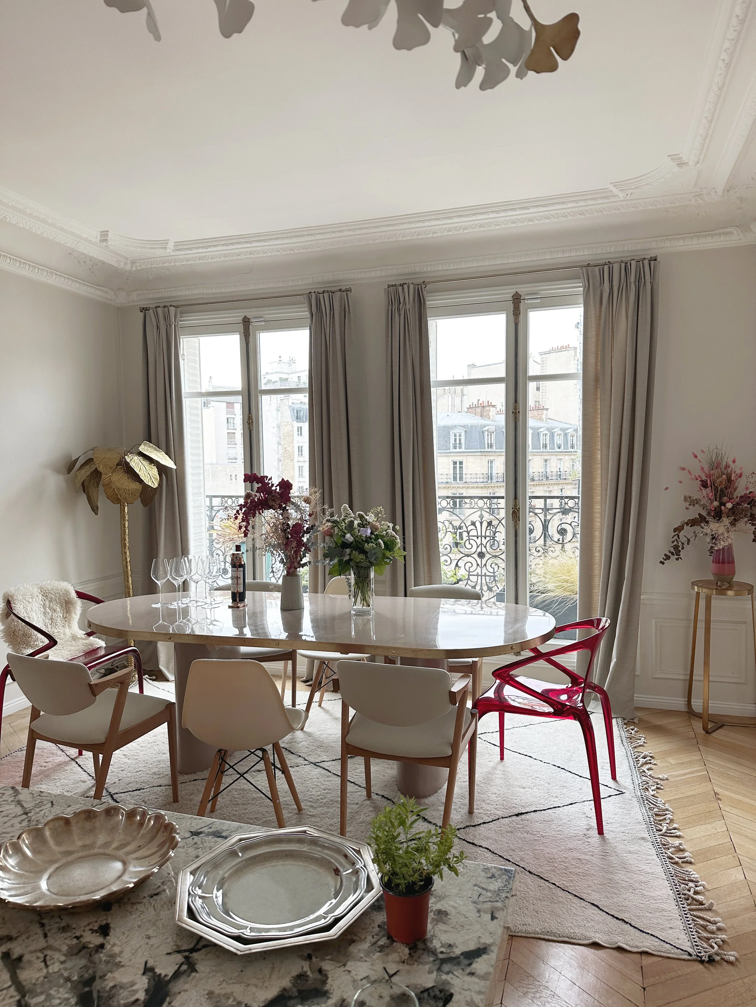 Une salle à manger lumineuse avec de grandes fenêtres et des rideaux beiges. Une table ronde en marbre blanc entourée de différentes chaises modernes, dont quelques chaises en plastique transparent rouge. Sur la table, plusieurs vases avec des fleurs