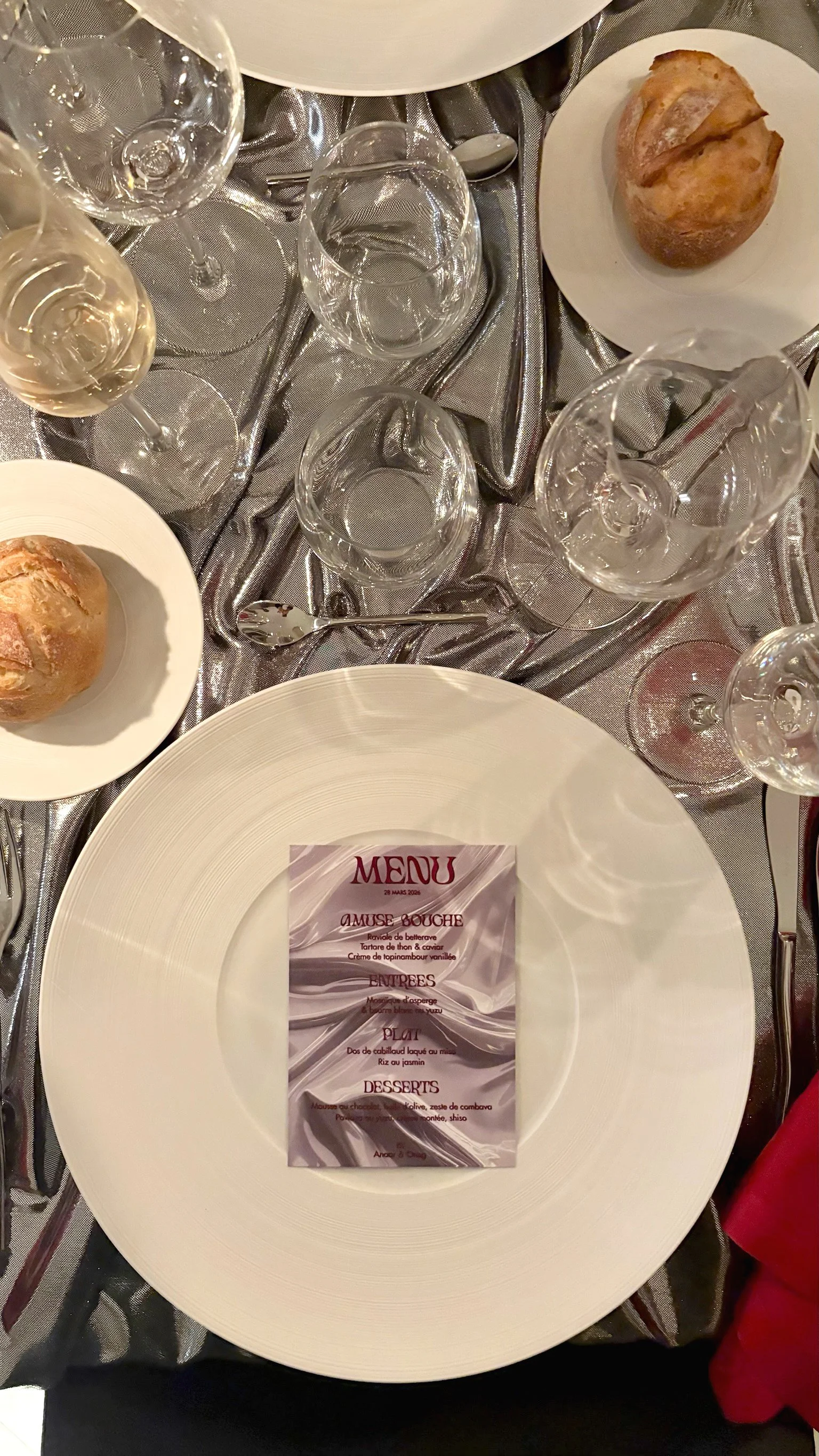 Une table de restaurant élégante avec des verres à vin, des assiettes, un pain, et un menu au centre. Le menu indique les plats et desserts disponibles.