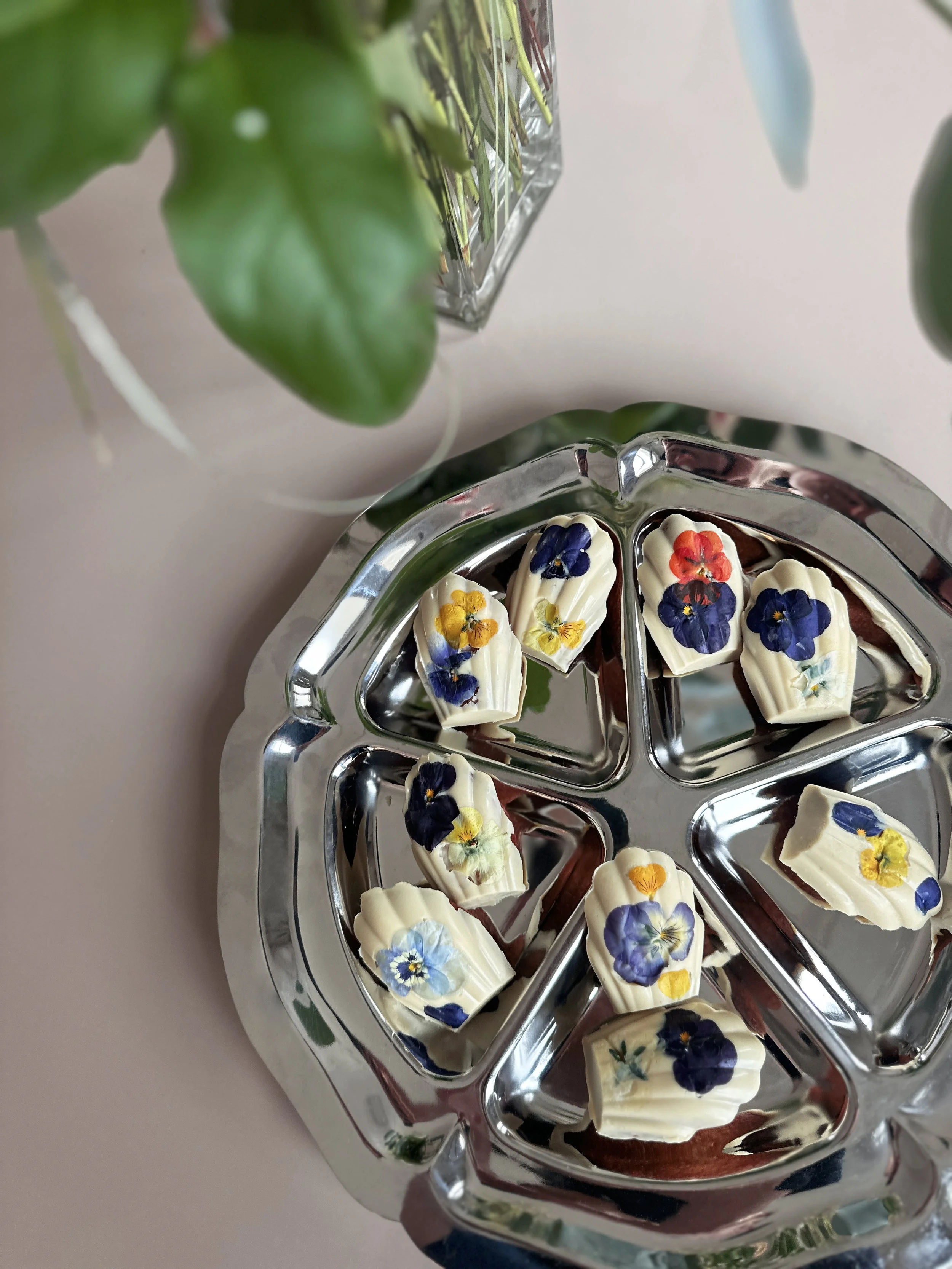 Plateau de madeleines en coque de chocolat blanc avec des fleurs comestibles violettes, jaunes, rouges et bleues, vue du dessus.