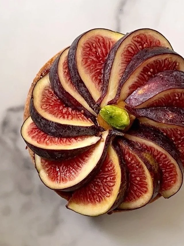 Tartelette de Figue coupée en fines tranches, formant un motif floral, avec une pistache verte au centre.