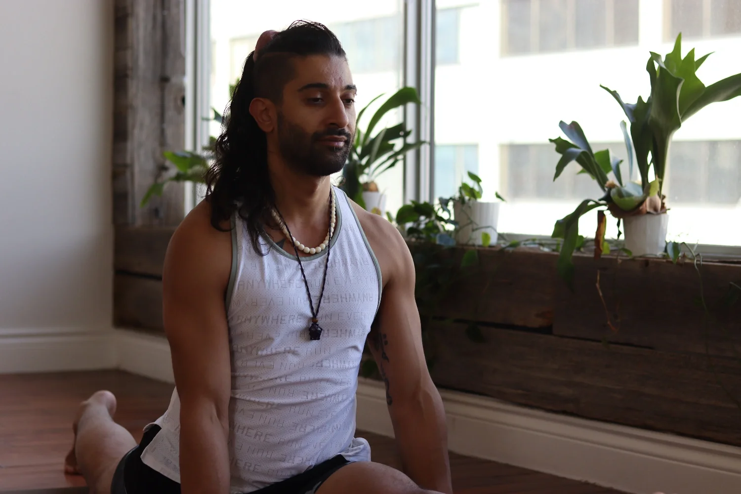 A person with long dark hair, wearing a white tank top and necklaces, practicing yoga or meditation in a room with large windows and potted plants.