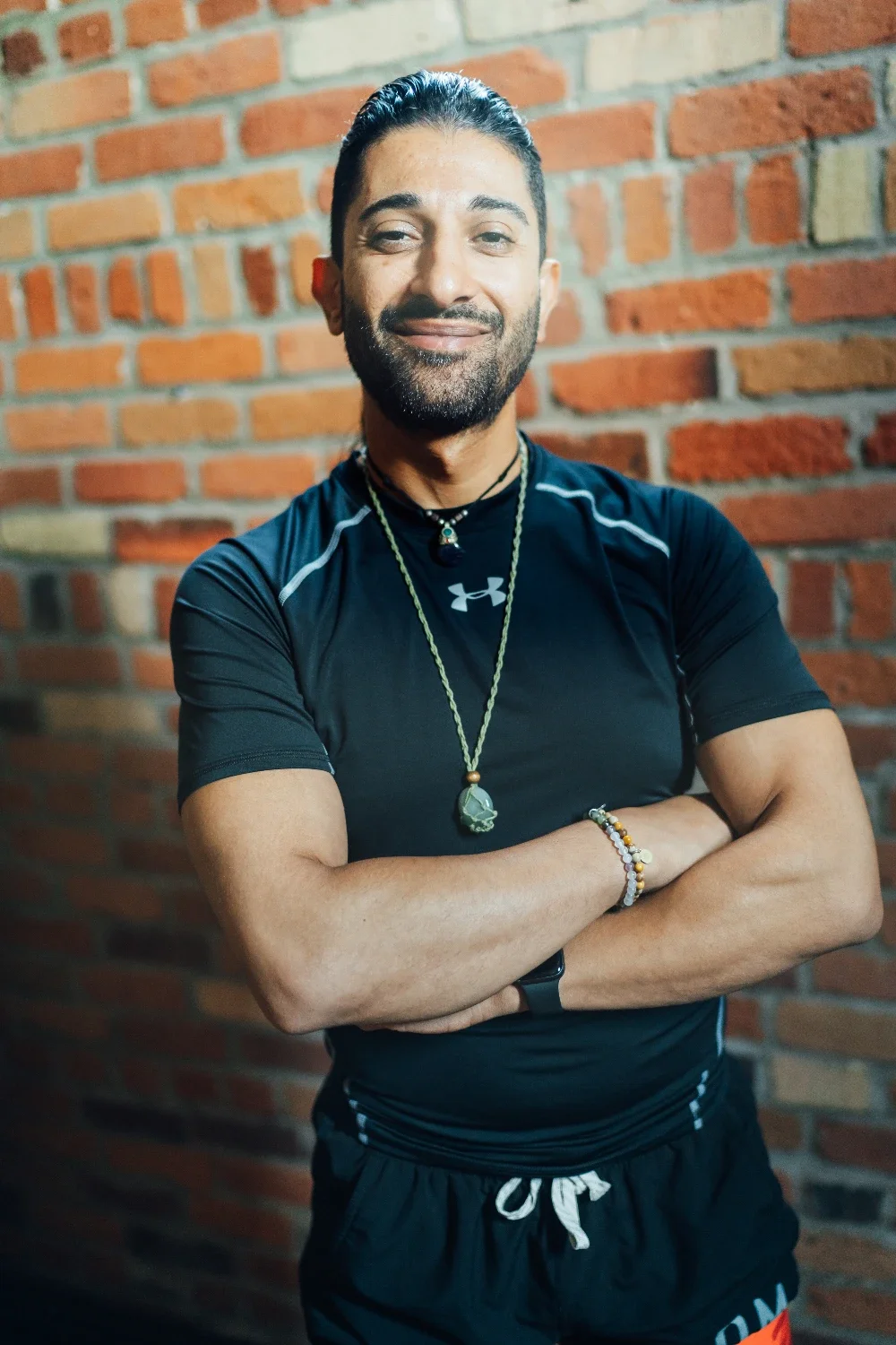 A smiling man with dark hair, beard, and tan skin, wearing a black athletic shirt, shorts, and various jewelry, standing confidently with arms crossed in front of a brick wall.