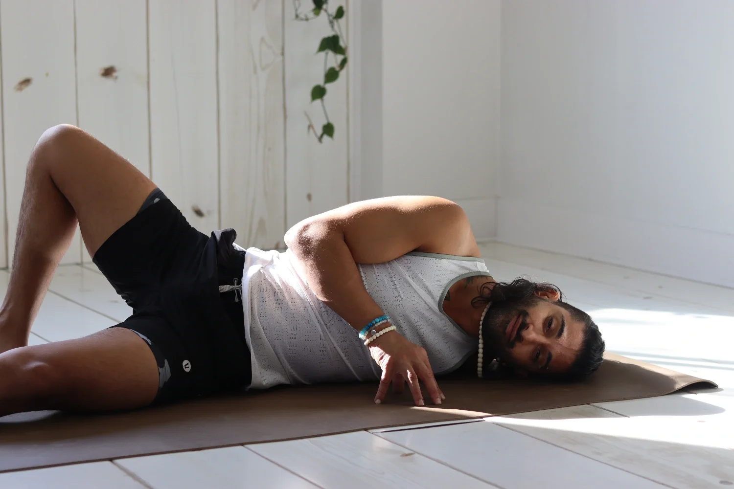 Man lying on his side on a yoga mat in a bright room with white wall and wooden floor, wearing a white tank top, black shorts, and bracelets, looking at the camera.