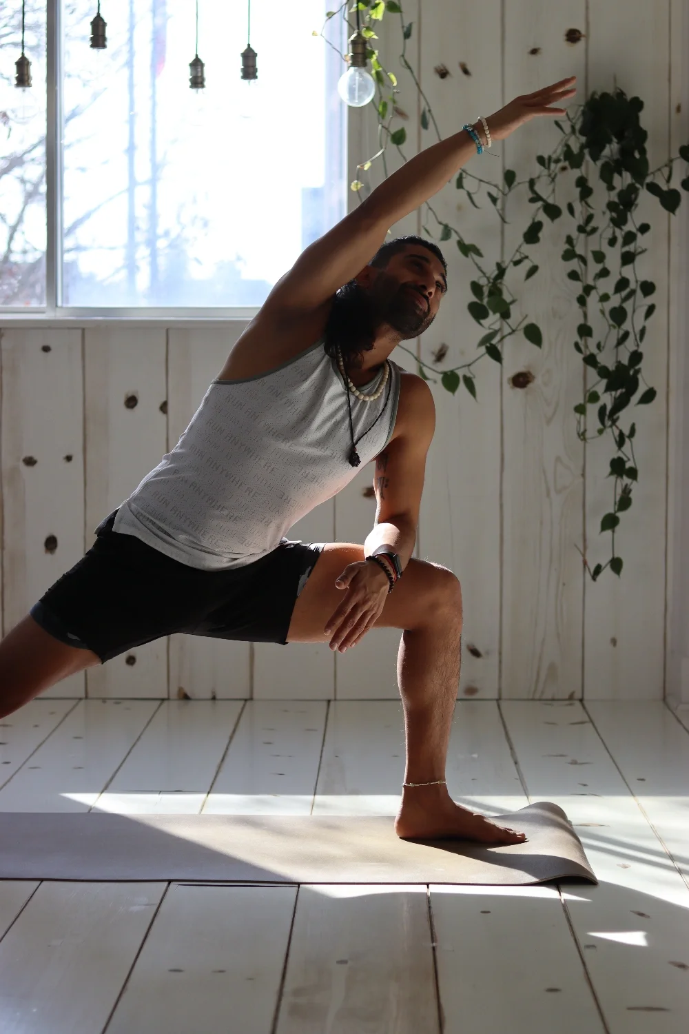 A man practicing yoga indoors, performing a lunge stretch with one arm extended overhead, on a yoga mat in a room with wooden walls and a large window.