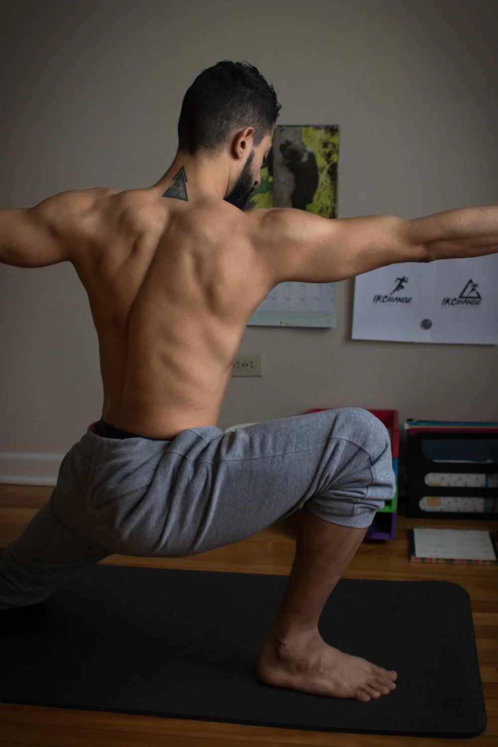 A shirtless man practicing yoga indoors, performing a low lunge pose with arms extended. He has a tattoo on his neck and is wearing gray sweatpants.