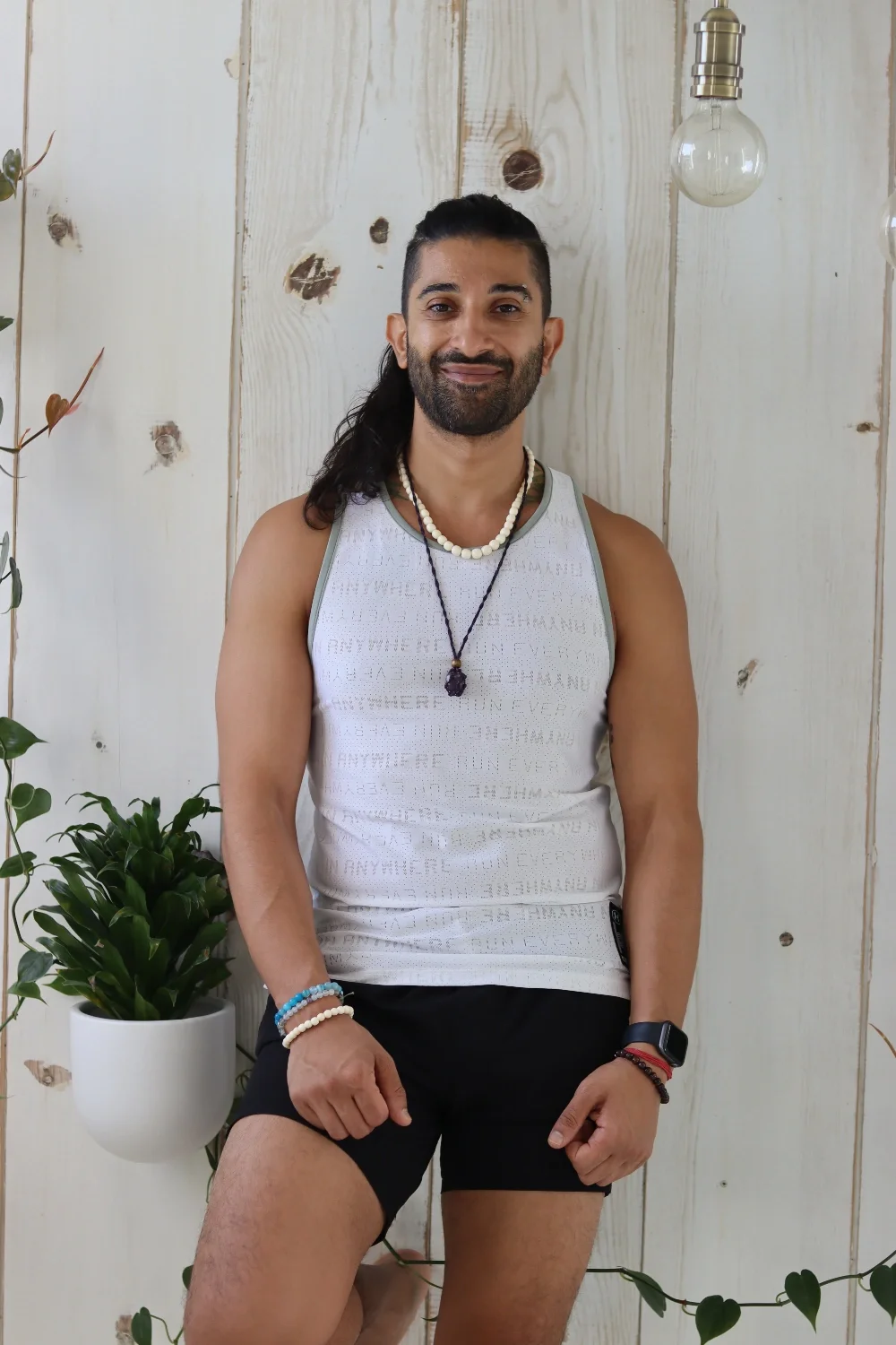 A smiling man with a beard and long dark hair tied back, wearing a white tank top with light green trim, black shorts, and multiple necklaces and bracelets, standing in front of a light wood-paneled wall with decorative plants and hanging light bulbs.