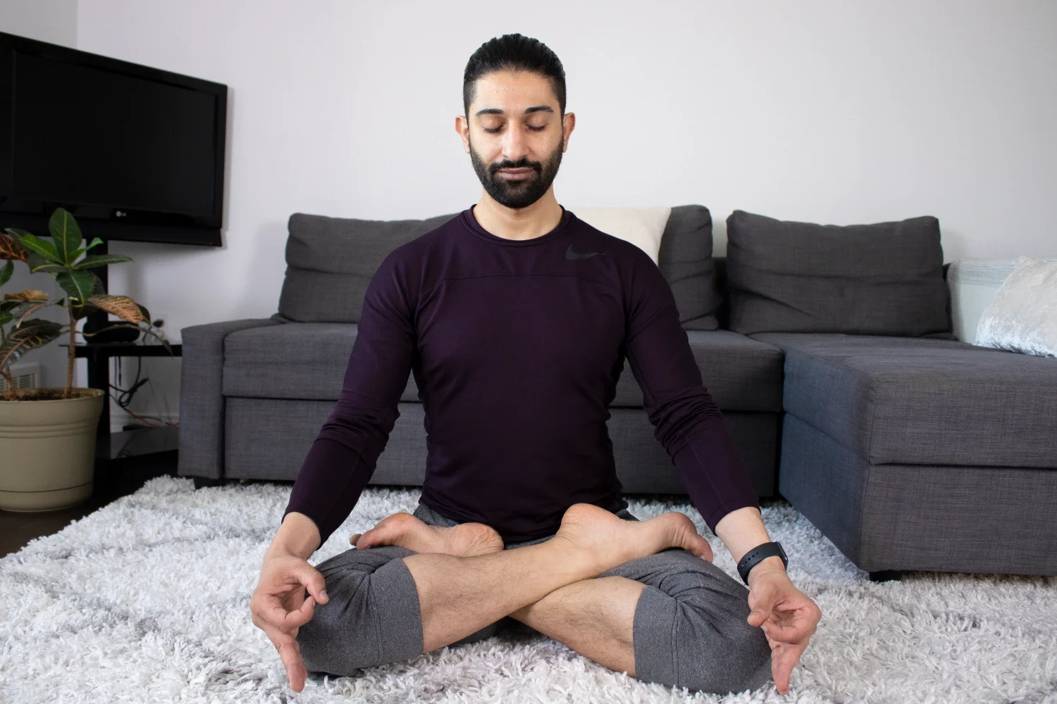 A man sitting cross-legged on a white rug in a living room, meditating with eyes closed, wearing a dark purple shirt and gray shorts.