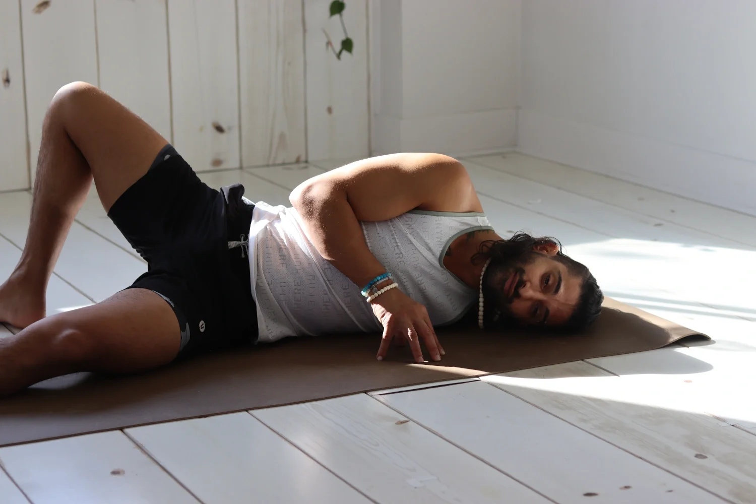 A man lying on a yoga mat on a wooden floor, in a relaxed pose, in a bright room with white wooden panel walls.