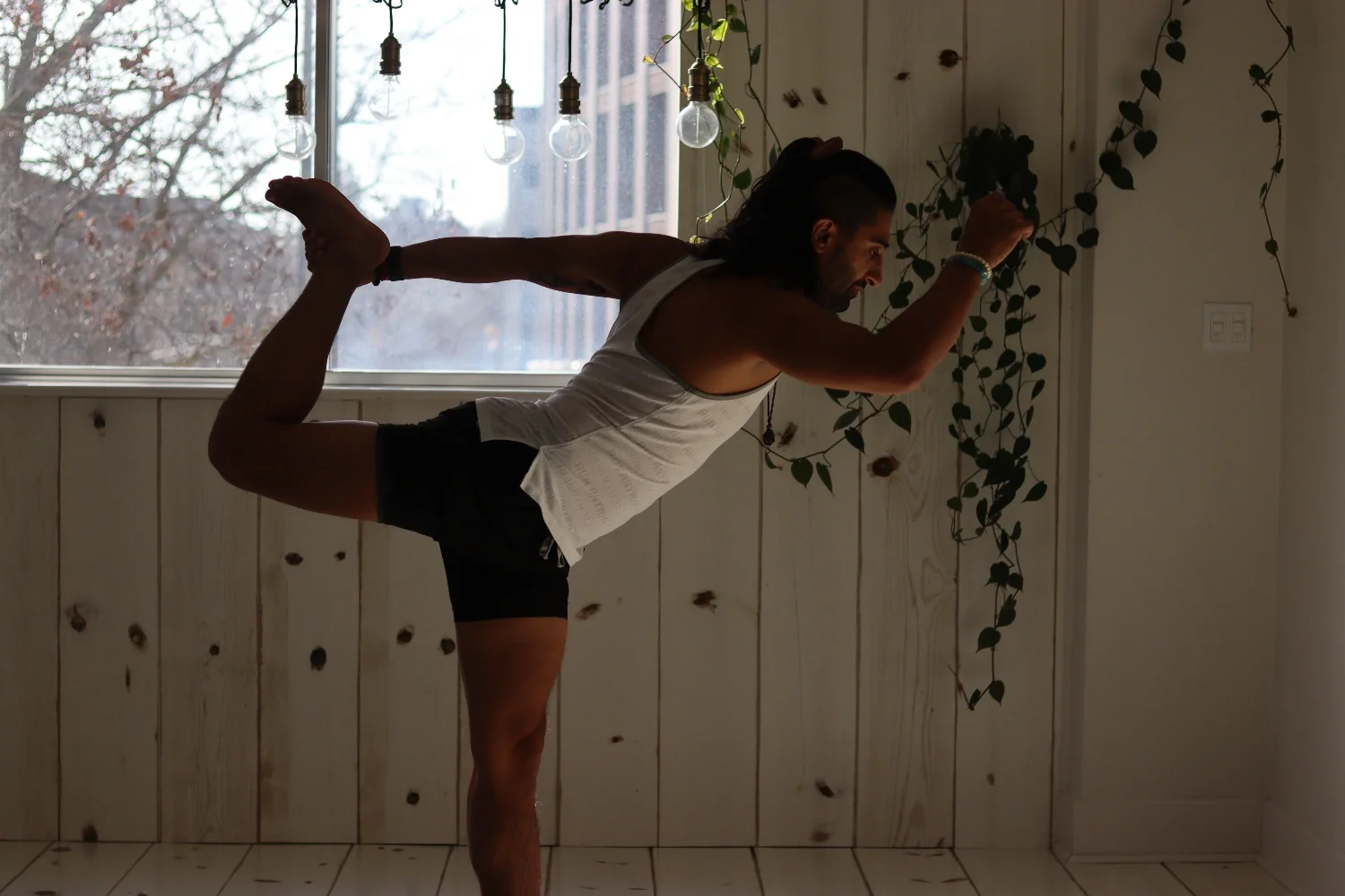 A man doing a yoga pose indoors near a window with hanging light bulbs and vines on the wall.