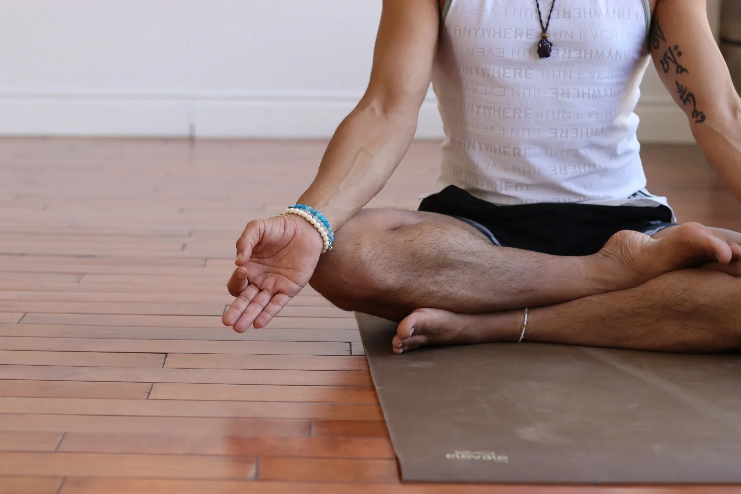 Person sitting cross-legged on a yoga mat, practicing meditation with one hand resting on their knee and the other hand in a mudra gesture. They are wearing a white tank top, black shorts, bracelets, and a necklace, with tattoos on their arms.