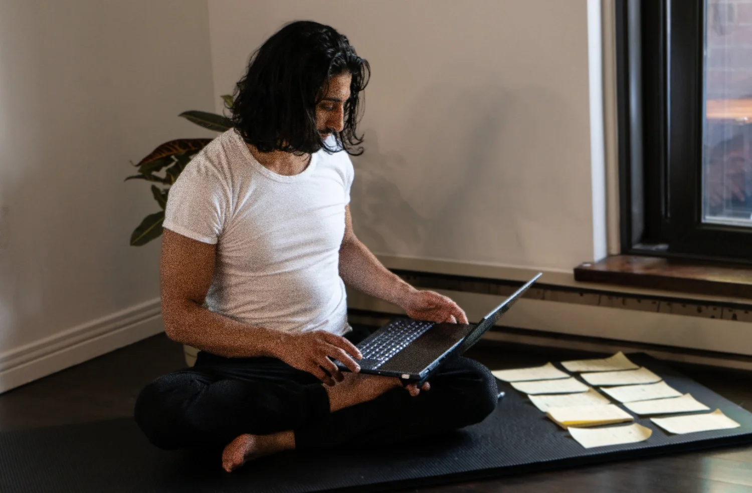 Person sitting cross-legged on a black yoga mat indoors, working on a laptop, with papers spread out on the floor nearby, near a window with natural light.