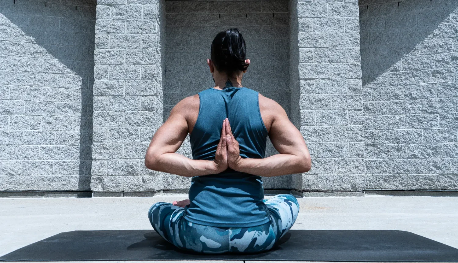 A woman practicing yoga outdoors, sitting cross-legged on a mat, wearing a teal sleeveless top and patterned leggings, with her hands in a prayer position behind her back.