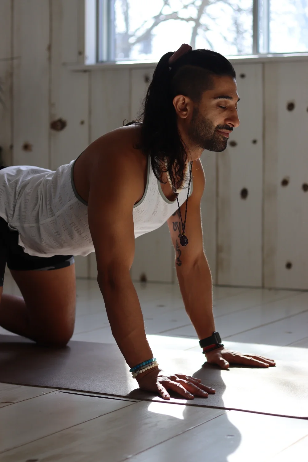 A man practicing yoga indoors in a plank position on a yoga mat, with sunlight coming through the window behind him.