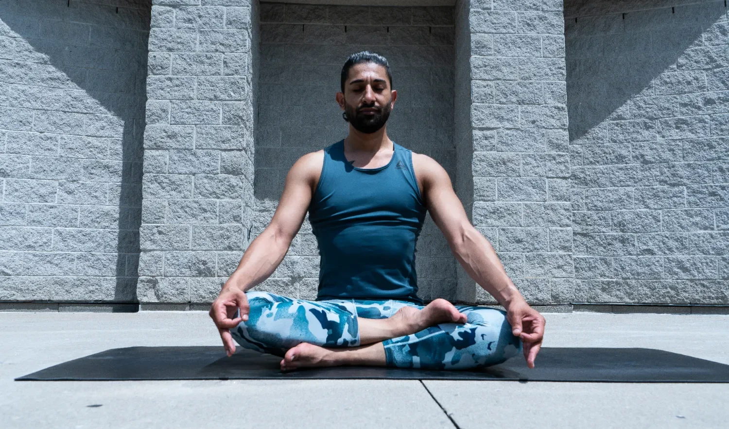 Man practicing yoga outdoors in a seated meditation pose with crossed legs and hands resting on knees, wearing a blue tank top and patterned blue leggings, against a gray brick wall.