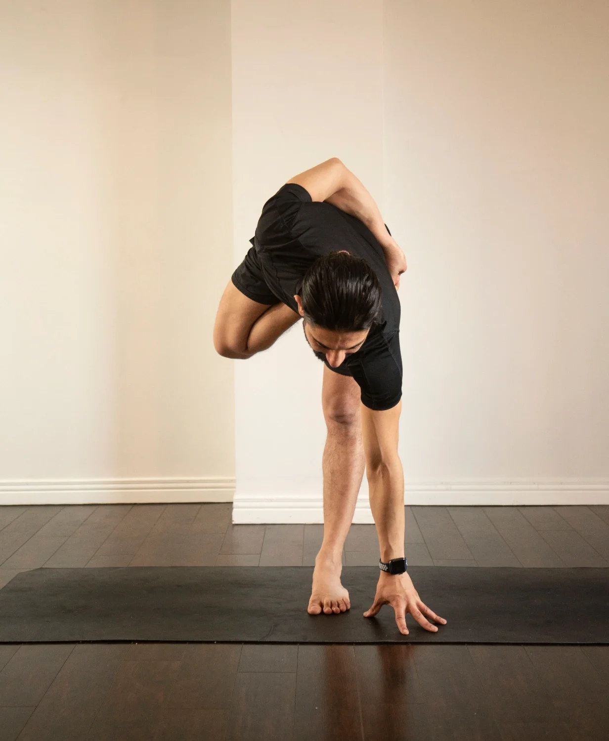 A person practicing yoga indoors, performing a forward fold pose with one hand on the ground and the other arm wrapped around their back.