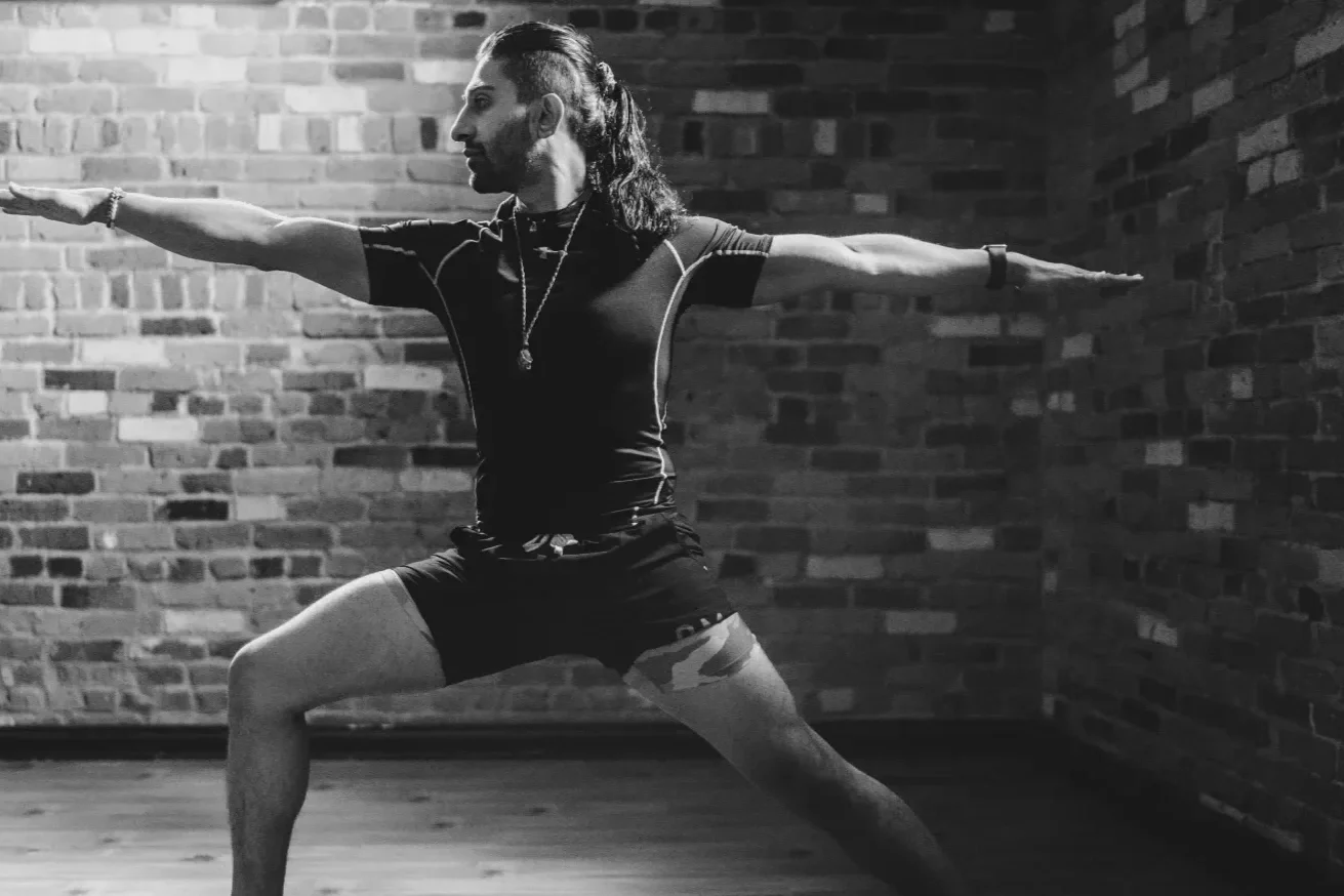 A man with long hair in a ponytail doing yoga in a brick-walled room, standing in warrior pose with arms extended.