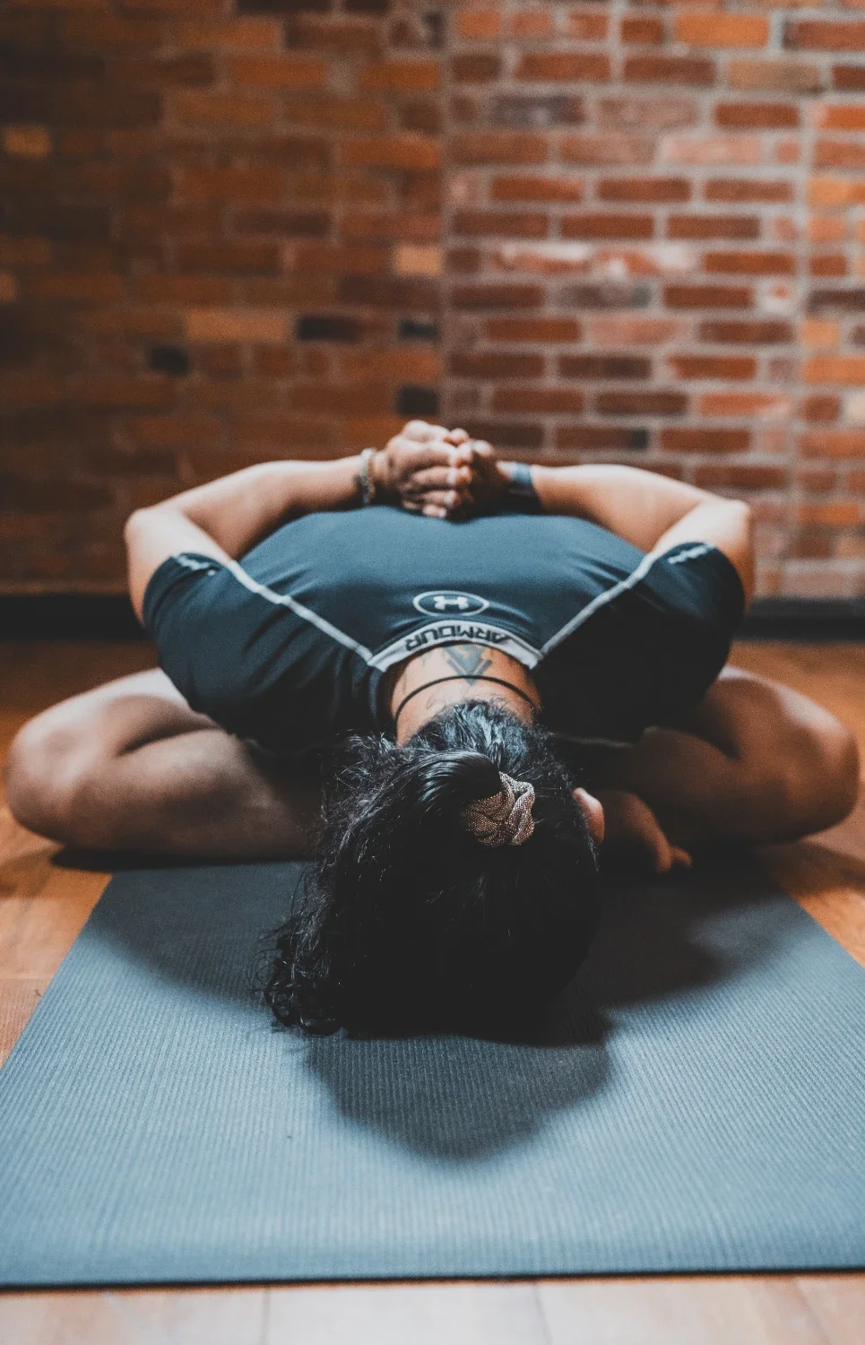 Person practicing yoga or meditation on a black mat against a brick wall, sitting in a cross-legged position with hands clasped behind their head.