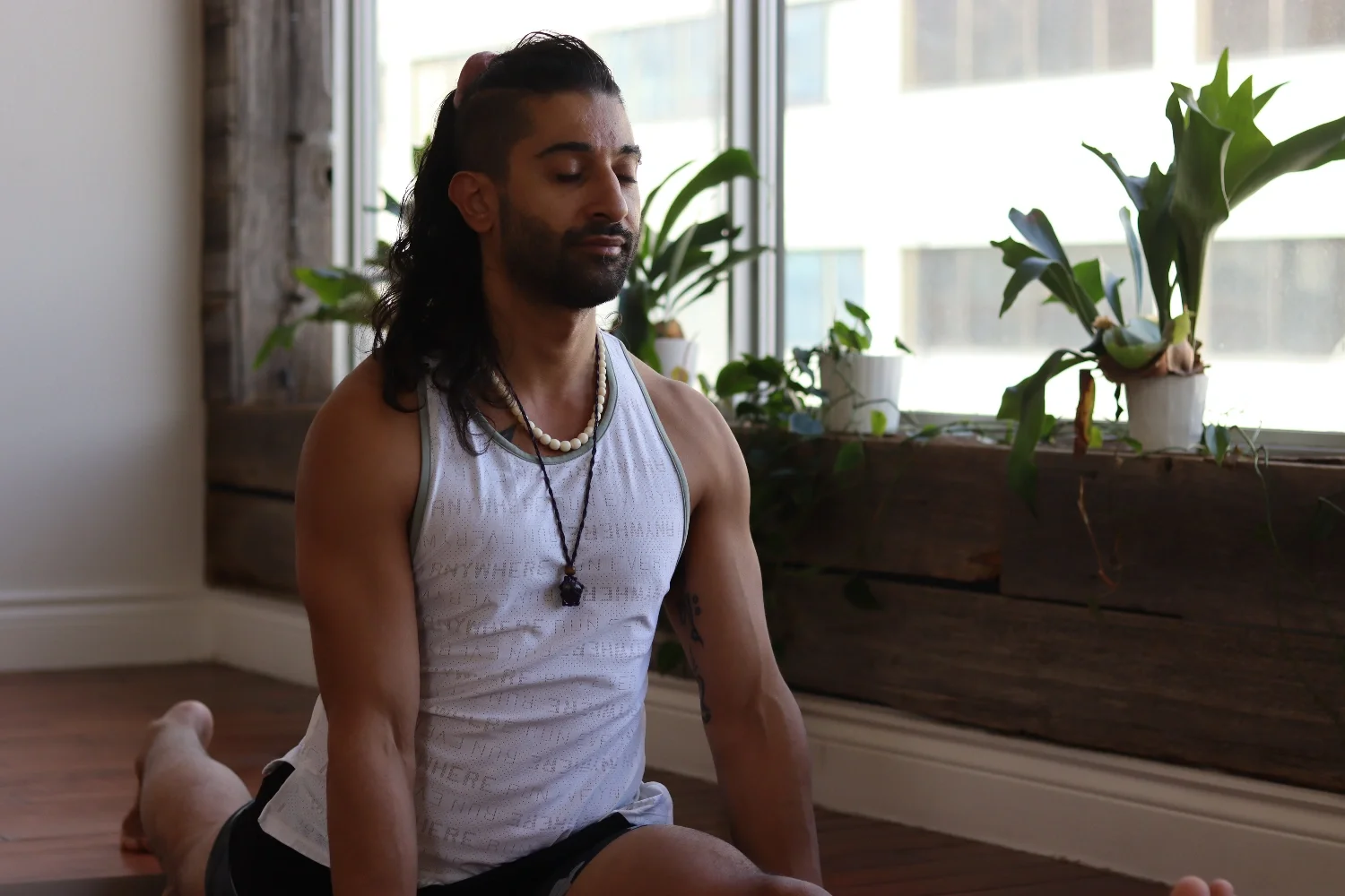 Man practicing yoga on a yoga mat indoors with plants on windowsill in background.