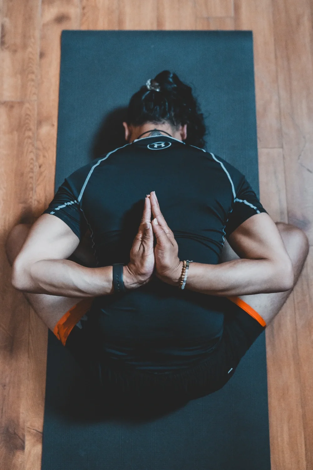 Person practicing yoga in a seated forward bend with hands in prayer position behind back, on a black yoga mat on wooden floor.