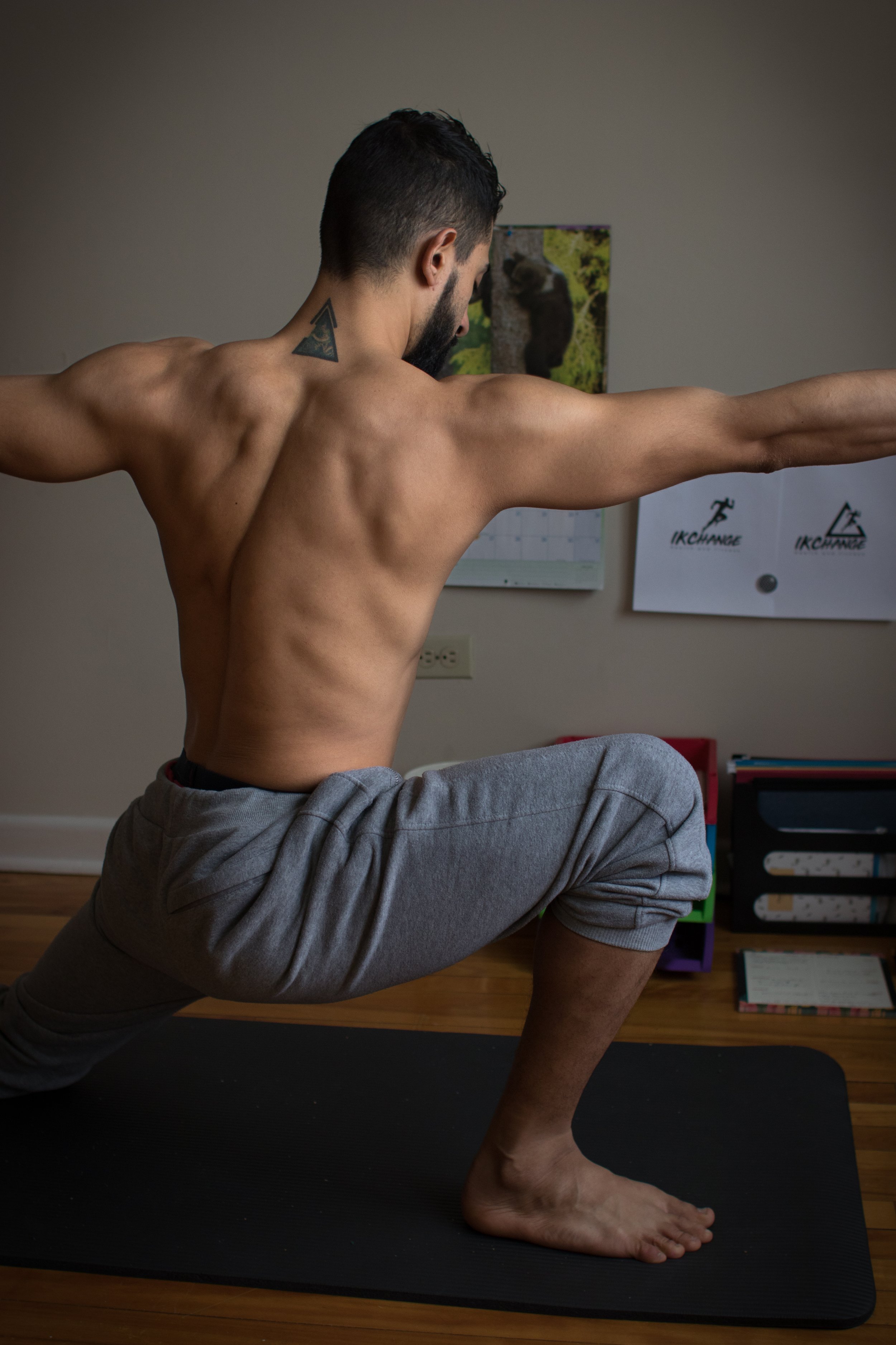 A shirtless man with a beard and short dark hair doing a yoga pose on a black mat, with his left knee on the floor, right foot flat, and left arm extended forward and right arm extended back, in a room with a calendar and a poster of a bear in the background.